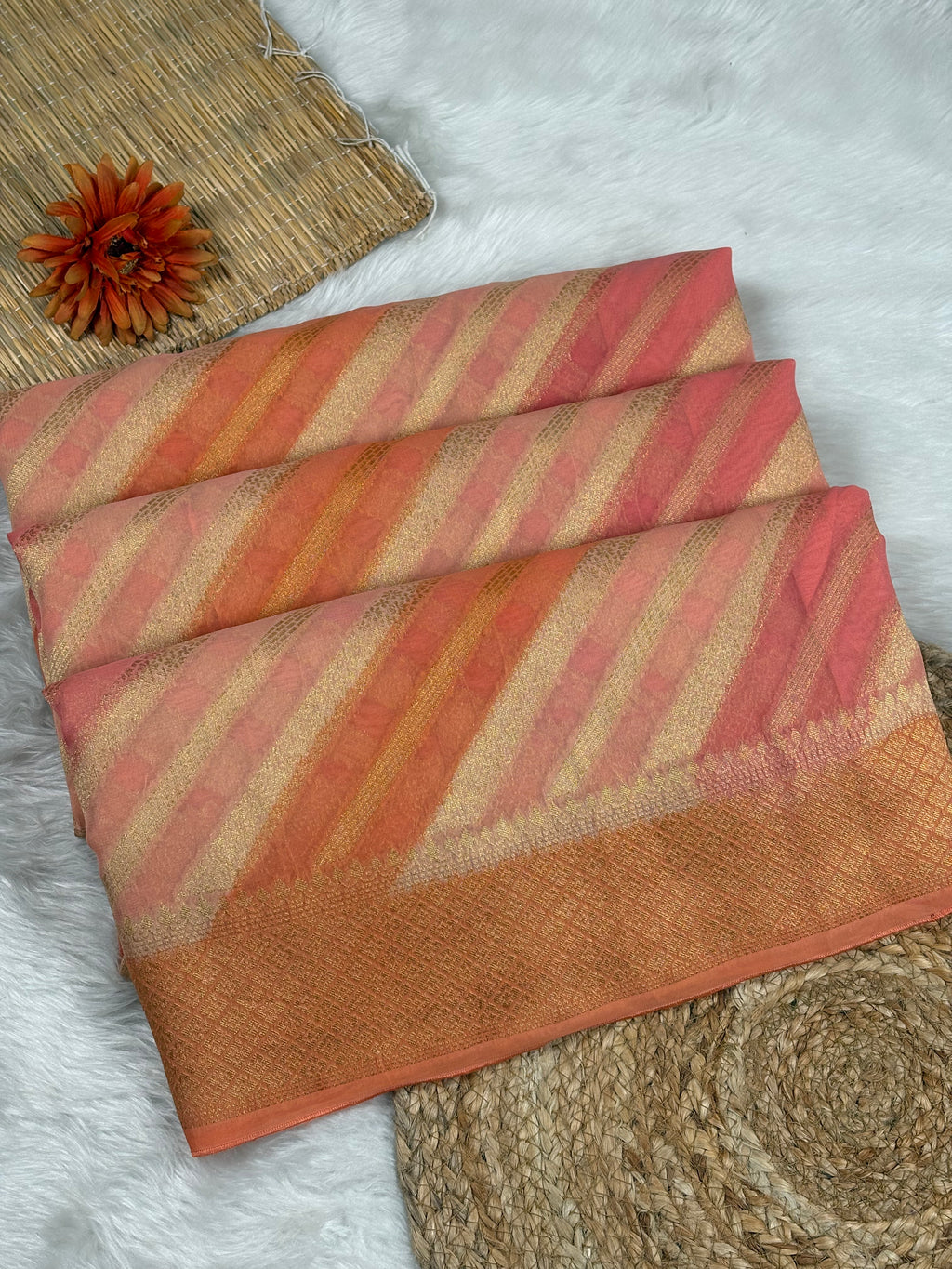 A folded striped saree, in shades of terracotta orange, dusty pink, and beige with a gold zari border, styled on a white faux fur surface with a woven mat and a decorative orange flower.