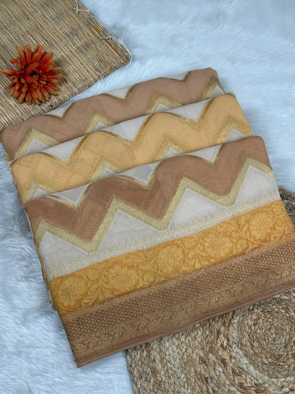 A folded patterned saree laid on a white faux-fur surface, with a woven mat and orange flower prop nearby. The product shows repeating chevron bands in beige, tan, cream, and mustard yellow with ornate woven detailing.