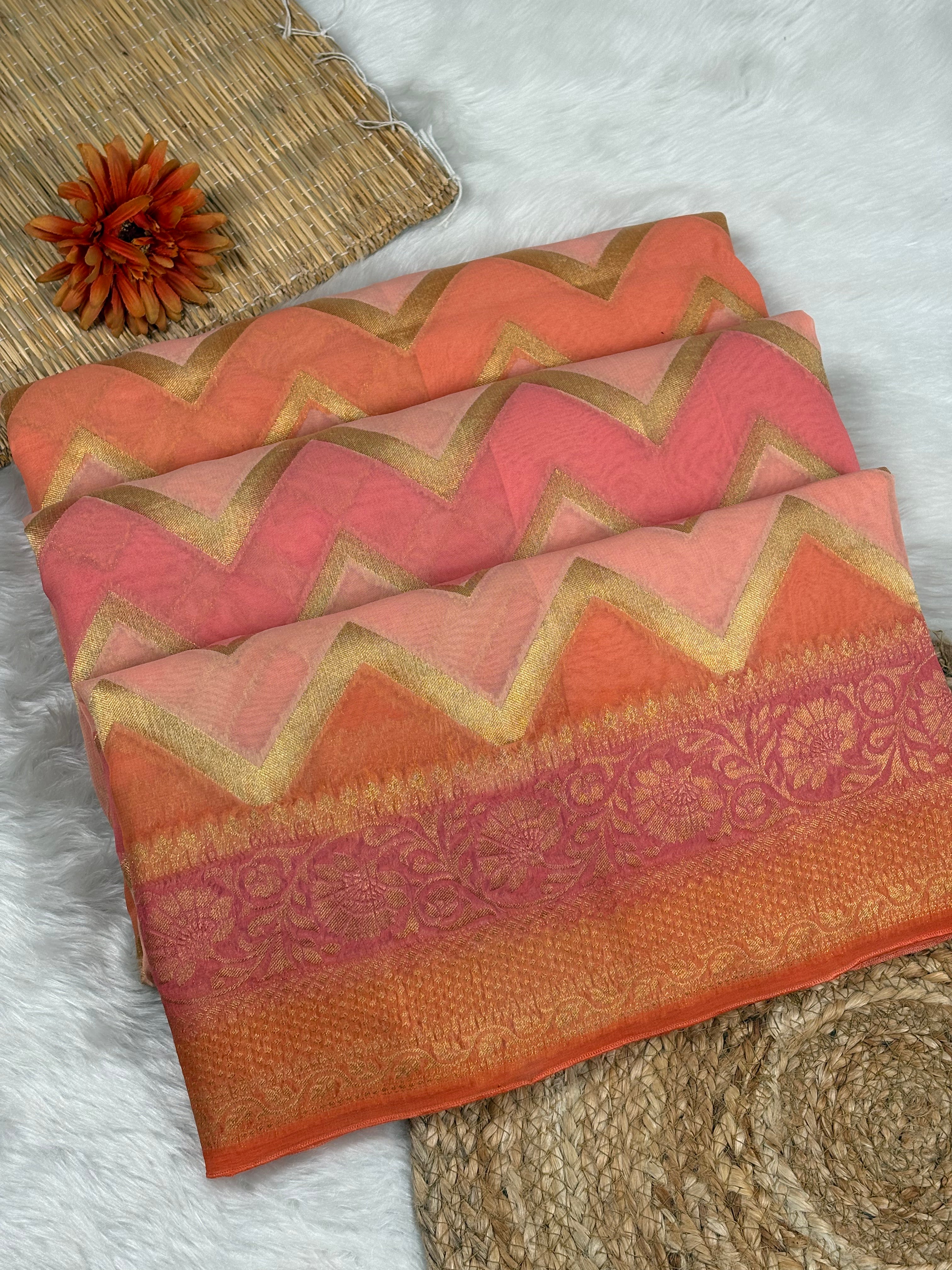 A folded orange and pink chevron-patterned saree with gold zari detailing, displayed on a white faux fur surface next to a woven mat and a dried orange flower.