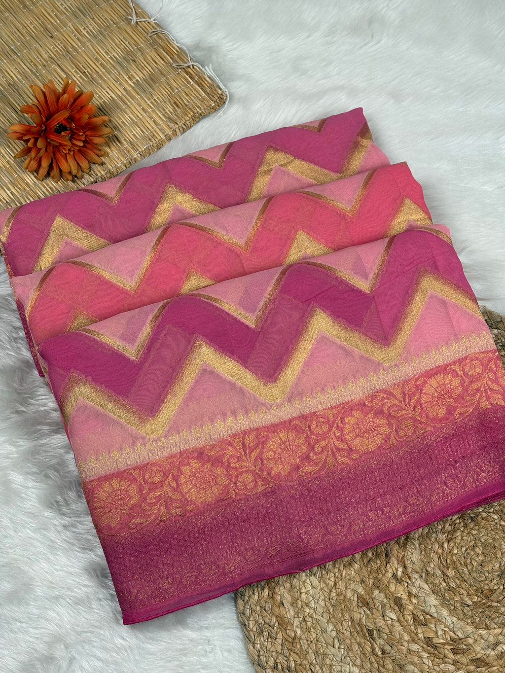 A folded pink and magenta chevron-patterned saree with gold zebra stripes and a floral border, displayed on a white faux-fur surface with woven props.