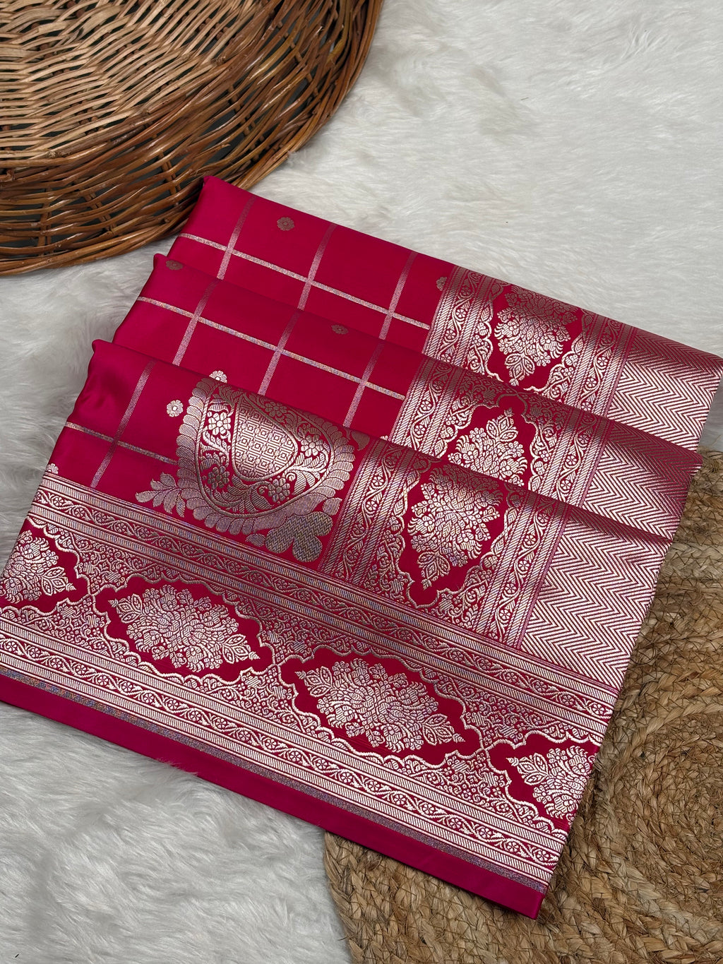 A folded red mashru katan Banarasi saree with intricate gold zari weaving, displayed on a white faux fur surface next to a woven basket.