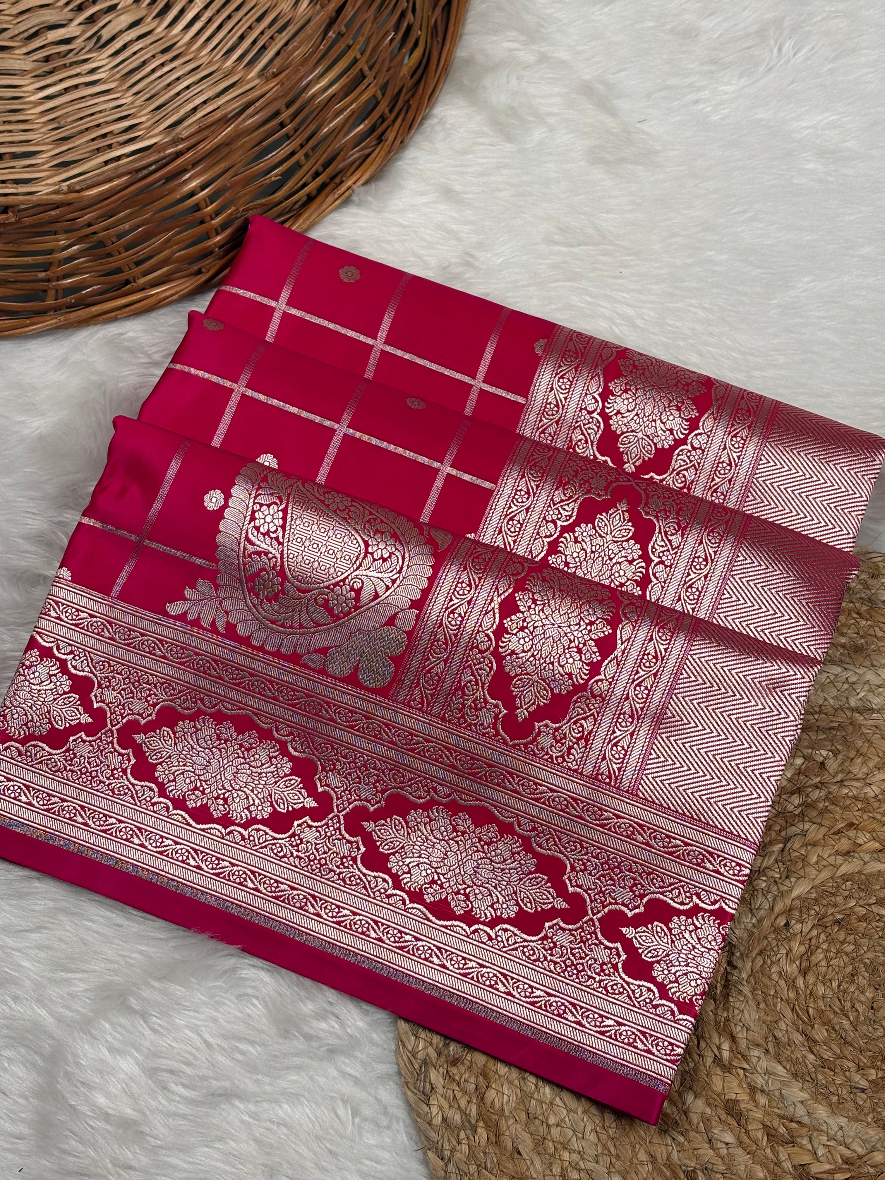 A folded red mashru katan Banarasi saree with intricate gold zari weaving, displayed on a white faux fur surface next to a woven basket.