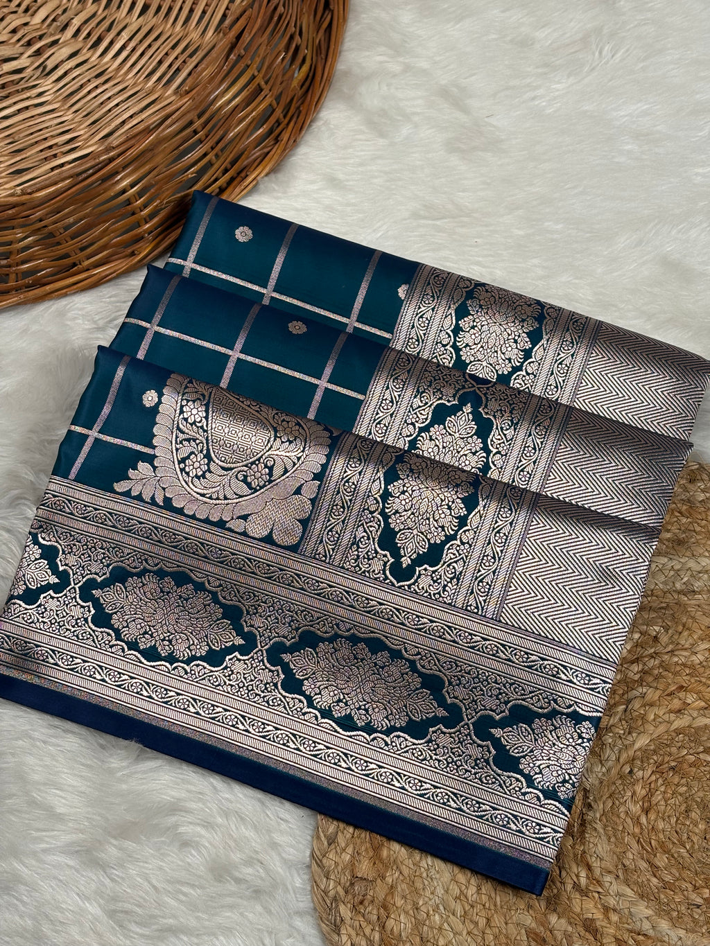 A folded dark blue saree with intricate gold zari woven patterns, laid out on a white faux fur surface next to a wicker basket.