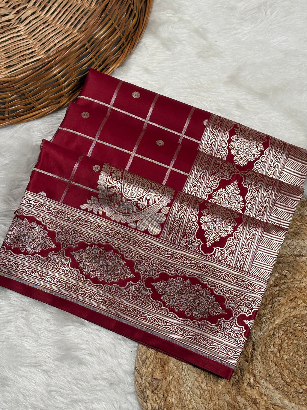 A folded deep red saree with intricate gold zari woven patterns, laid out on a white faux fur surface next to a wicker basket.