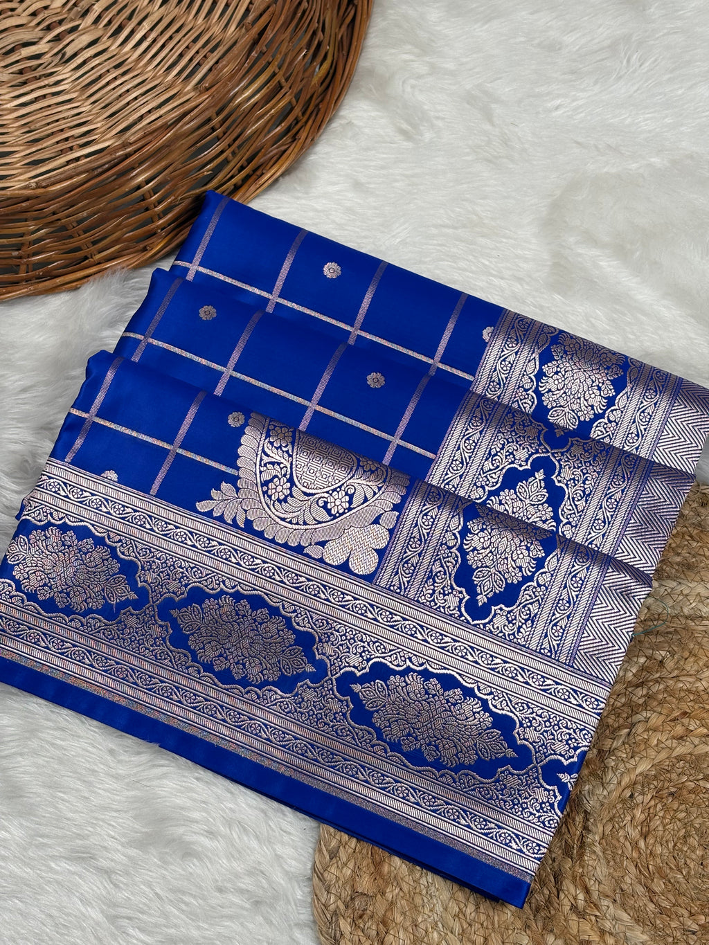 A royal blue mashru silk saree laid flat on a white faux fur surface, styled with a woven wicker basket and a beige textured mat.