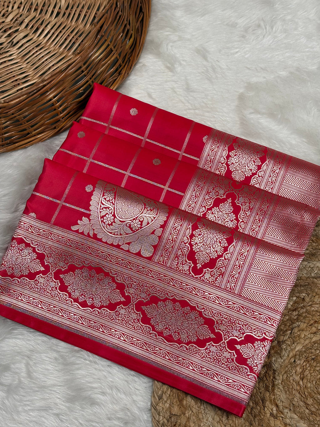 A folded red Banarasi silk saree with rose gold zari weaving, displayed on a white faux fur surface next to a woven wicker basket.