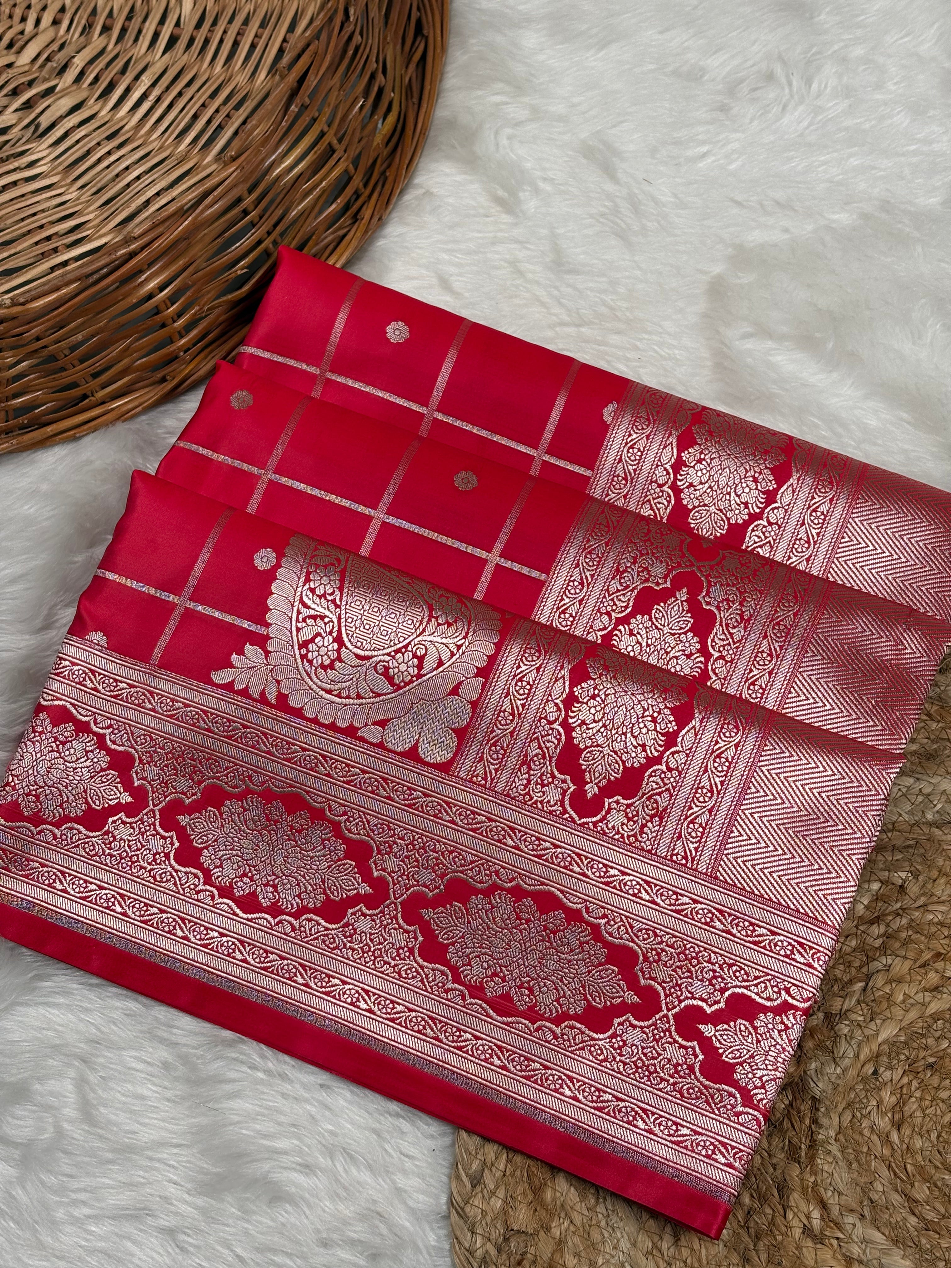 A folded red Banarasi silk saree with rose gold zari weaving, displayed on a white faux fur surface next to a woven wicker basket.