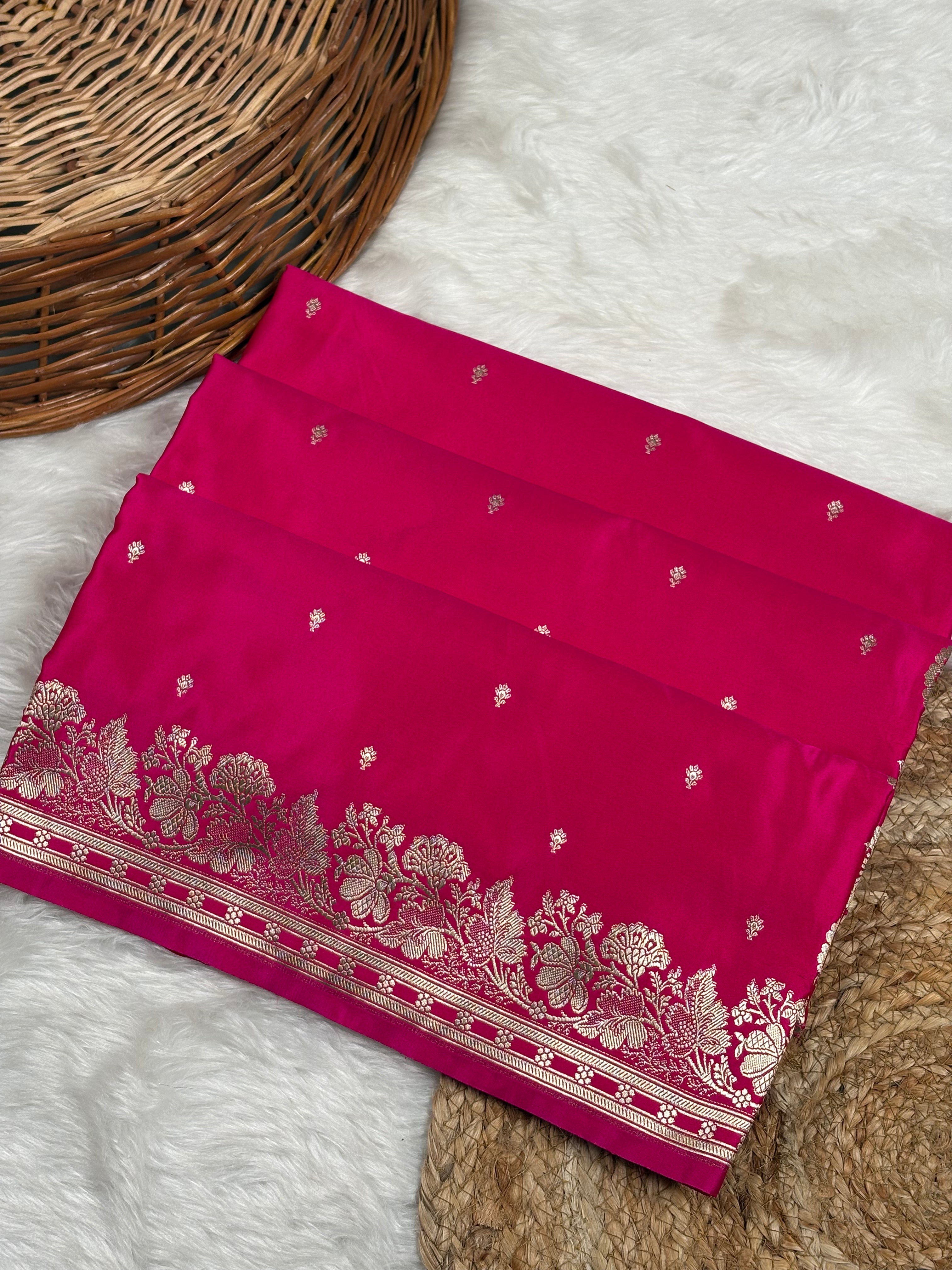 A folded hot pink saree with a wide gold zari floral border and small gold buti motifs, displayed on a white faux fur rug next to a wicker basket.