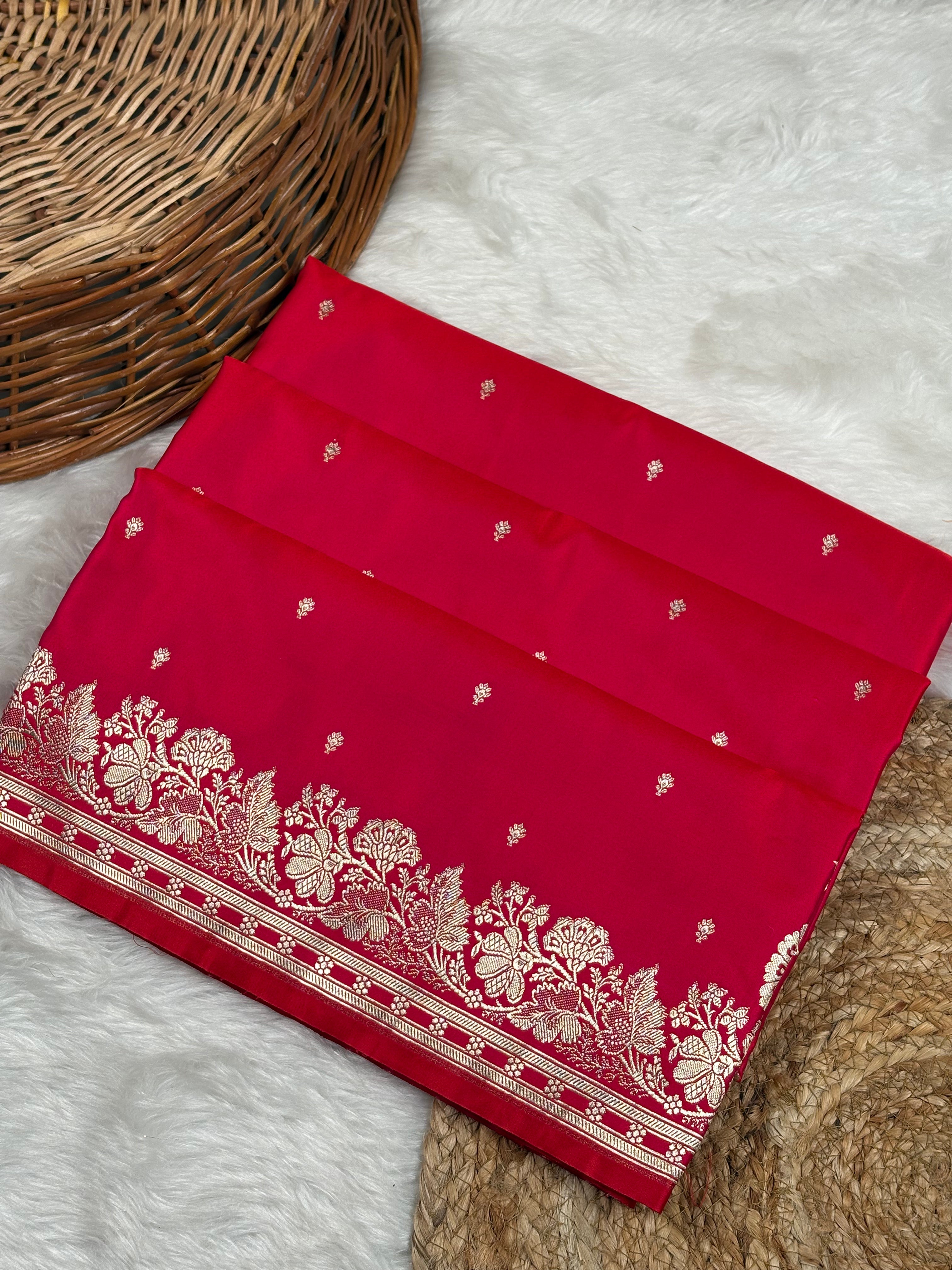 A folded red saree, featuring intricate gold zari floral border and small gold zari motifs, displayed on a white faux fur surface next to a wicker basket.