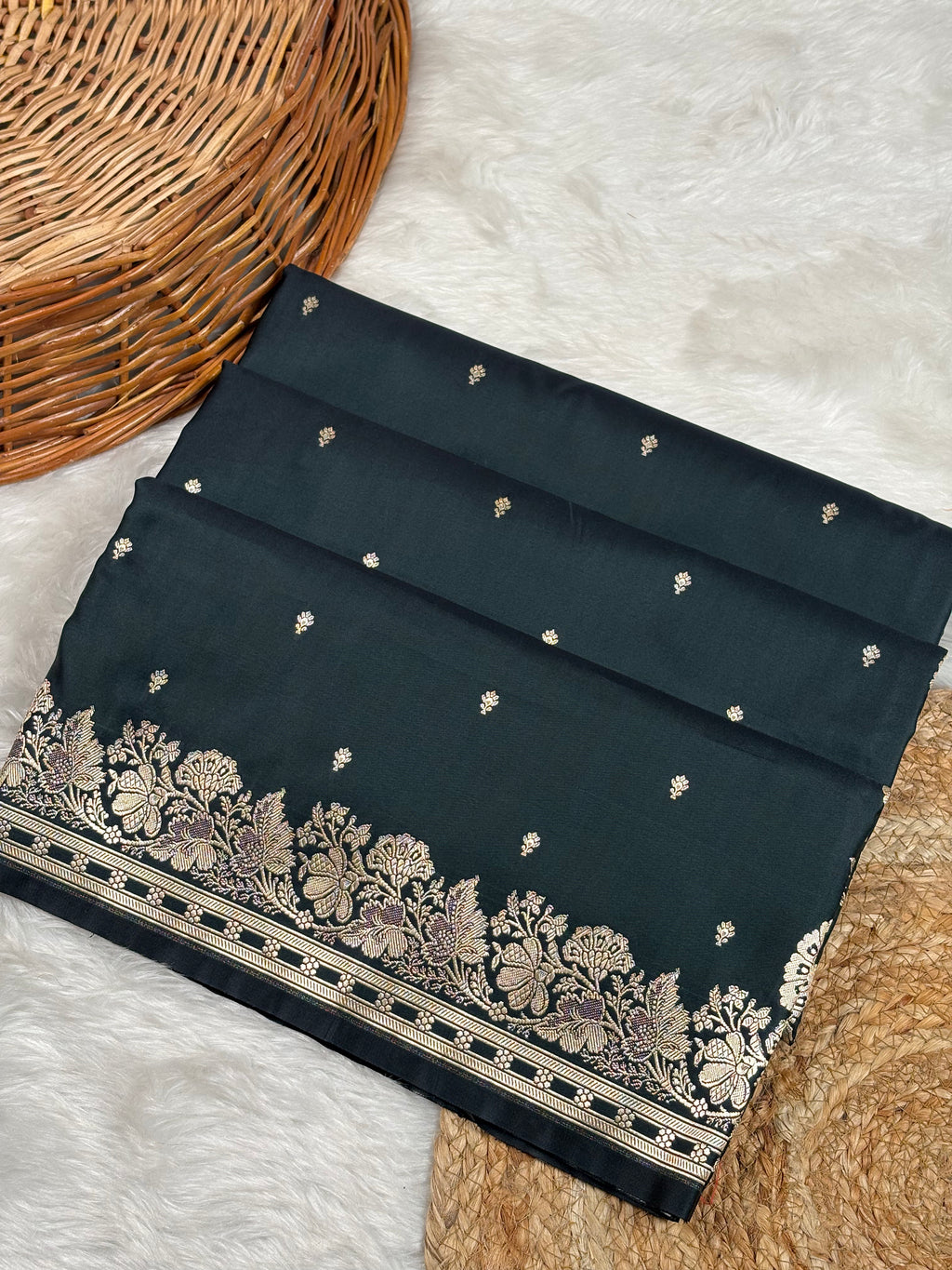 A folded dark black saree with small scattered motifs and a wide ornate floral border in gold zari weaving, styled on a white faux-fur surface with a wicker basket corner visible.