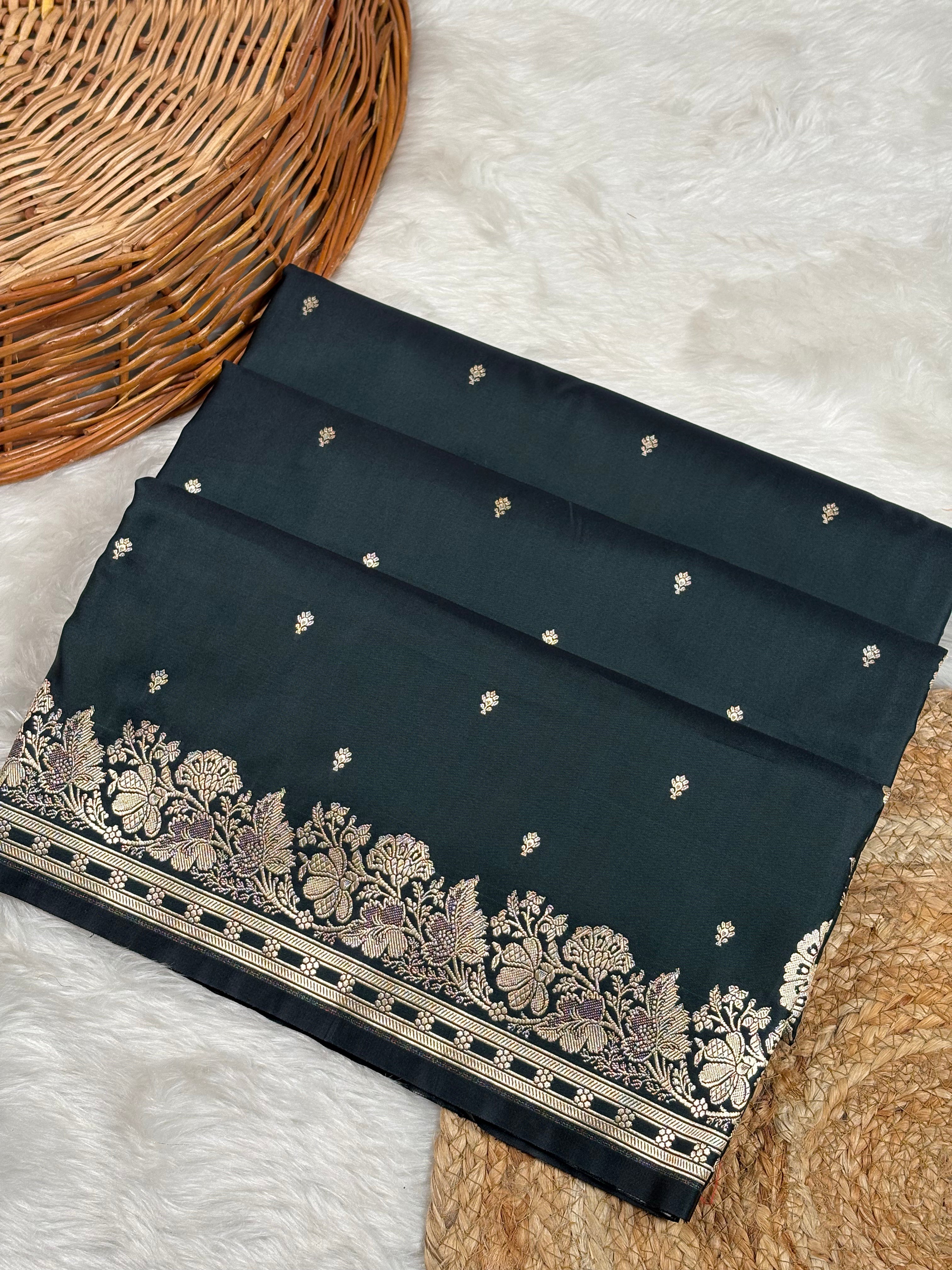 A folded dark black saree with small scattered motifs and a wide ornate floral border in gold zari weaving, styled on a white faux-fur surface with a wicker basket corner visible.