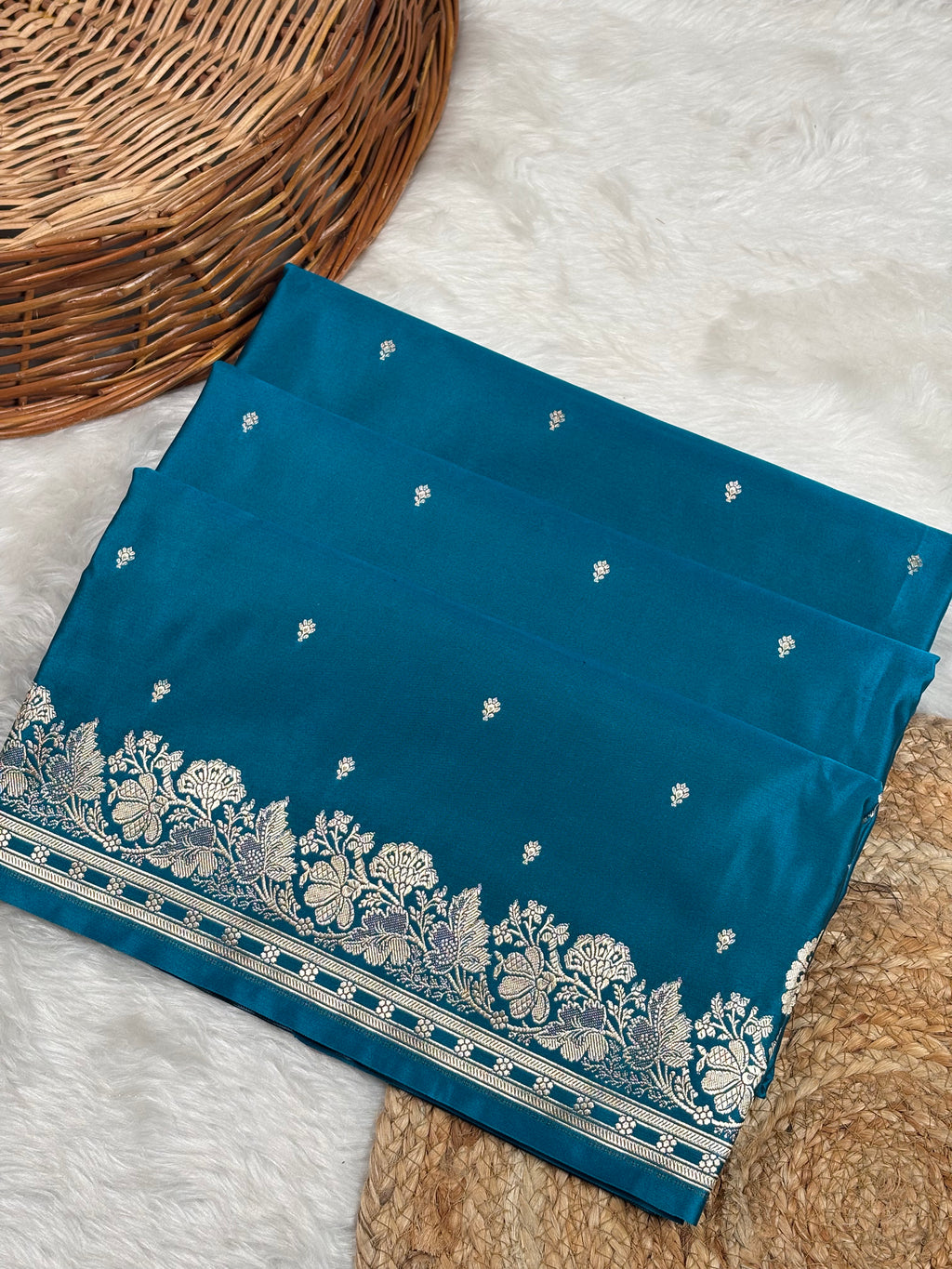 A folded teal blue saree, featuring a detailed gold zari floral border and small gold zari buti motifs weaving, displayed on a white faux fur rug next to a wicker basket.
