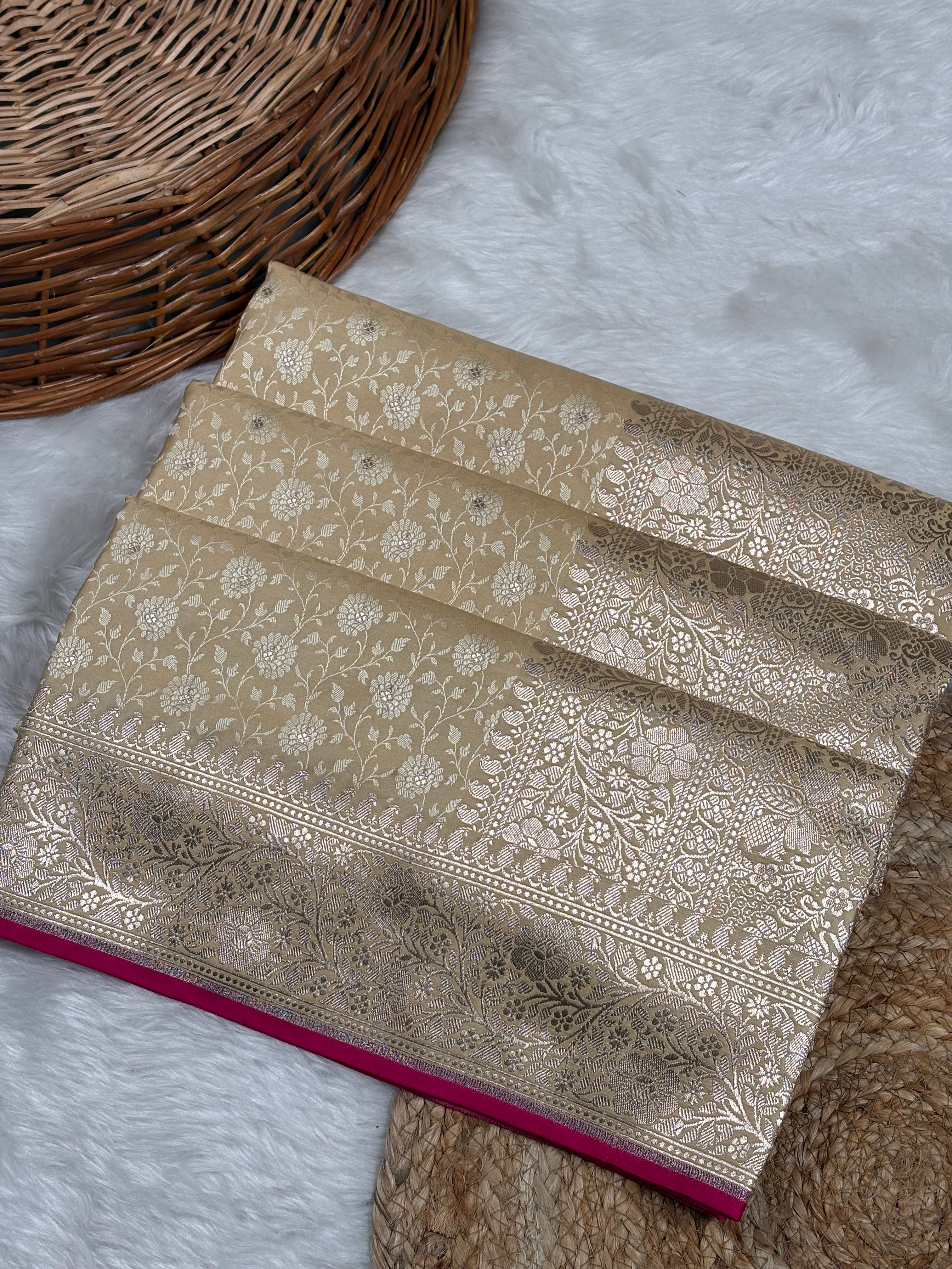 A folded beige and gold brocade saree with intricate floral patterns border, displayed on a white faux fur surface next to a wicker basket.