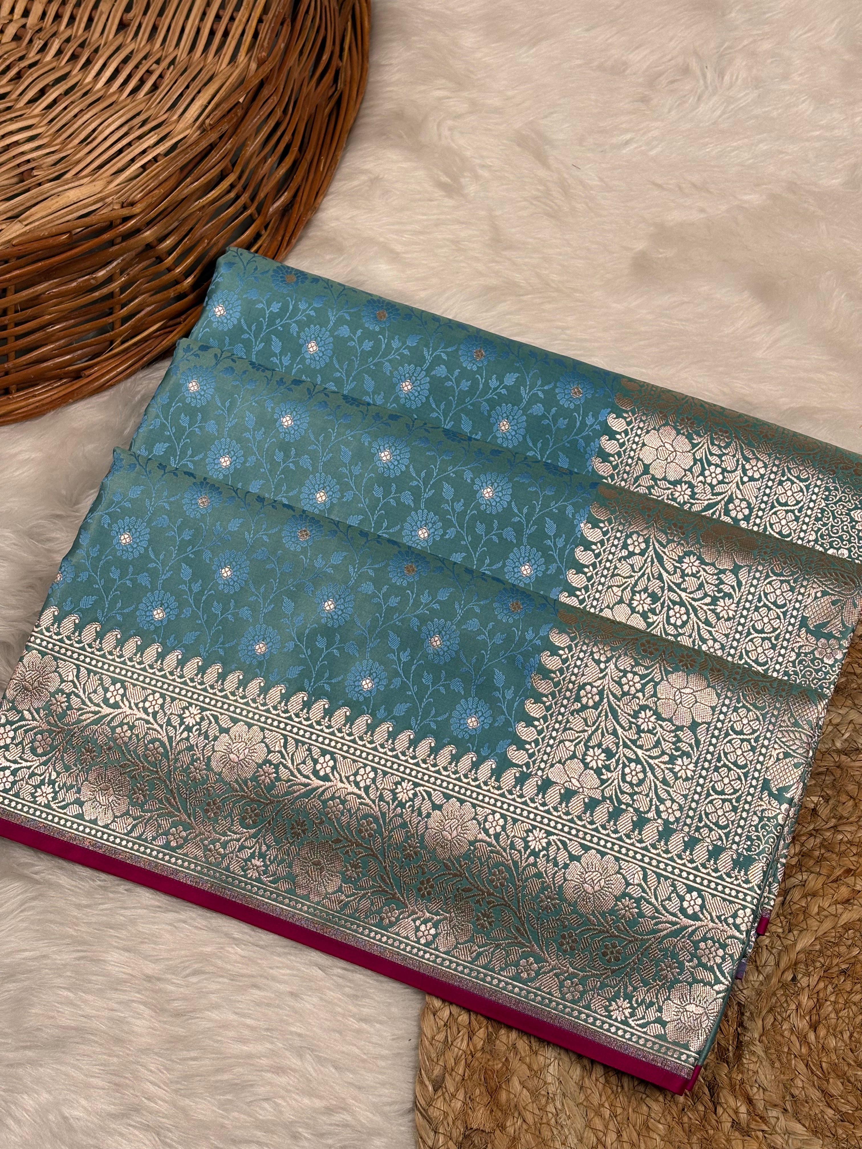 A folded teal blue Banarasi silk saree with intricate gold zari floral patterns and a wide border, displayed on a white faux fur rug next to a wicker basket.
