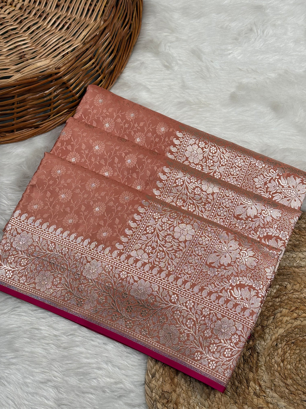 A folded terracotta pink Banarasi silk saree with intricate gold zari floral weaving, displayed on a white faux fur rug next to a woven wicker basket.