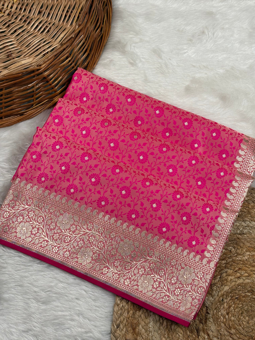 A folded pink Banarasi silk saree with intricate gold zari floral weaving and a wide ornate border, displayed on a white faux fur rug next to a wicker basket.