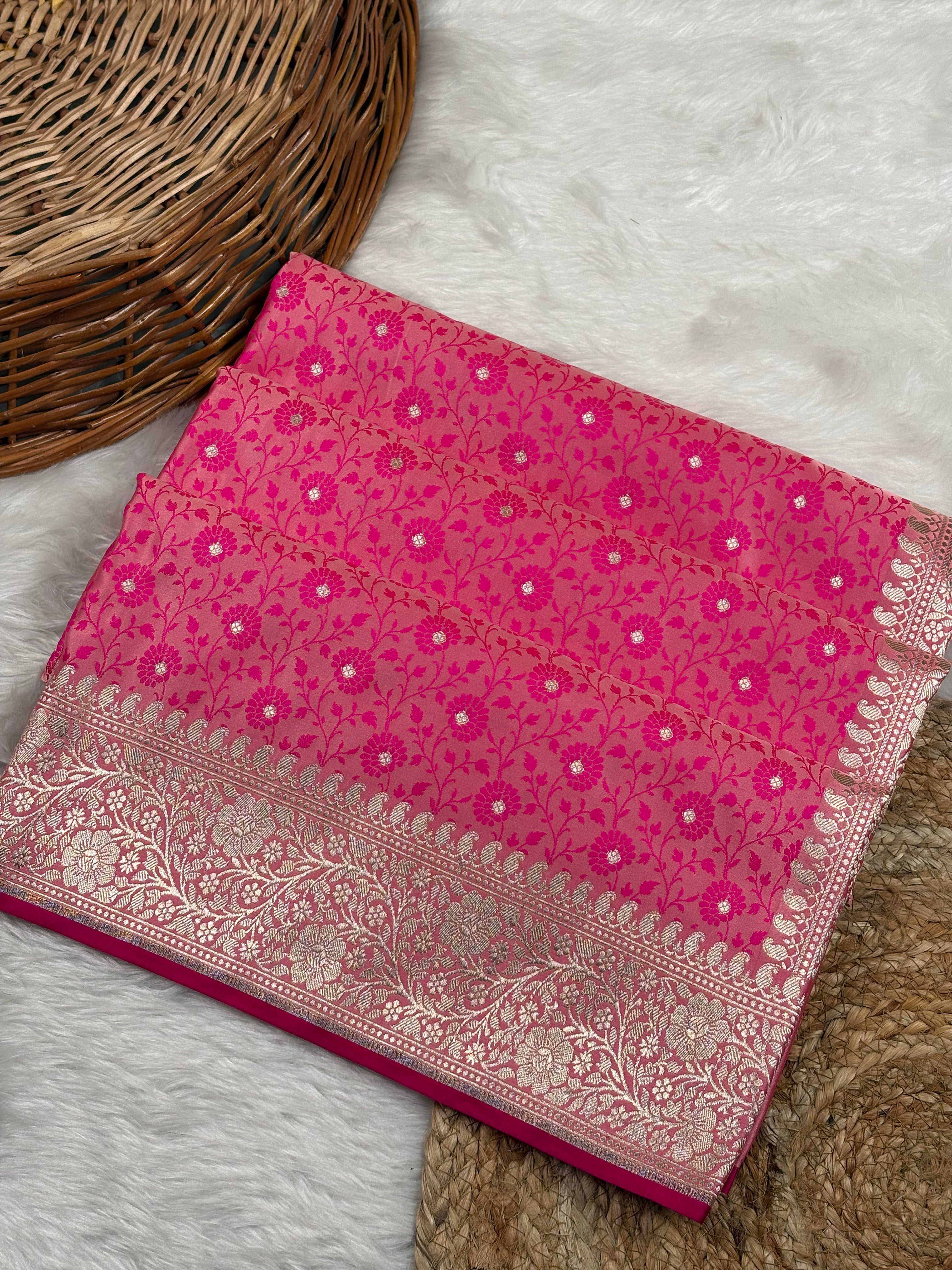 A folded pink Banarasi silk saree with intricate gold zari floral weaving and a wide ornate border, displayed on a white faux fur rug next to a wicker basket.