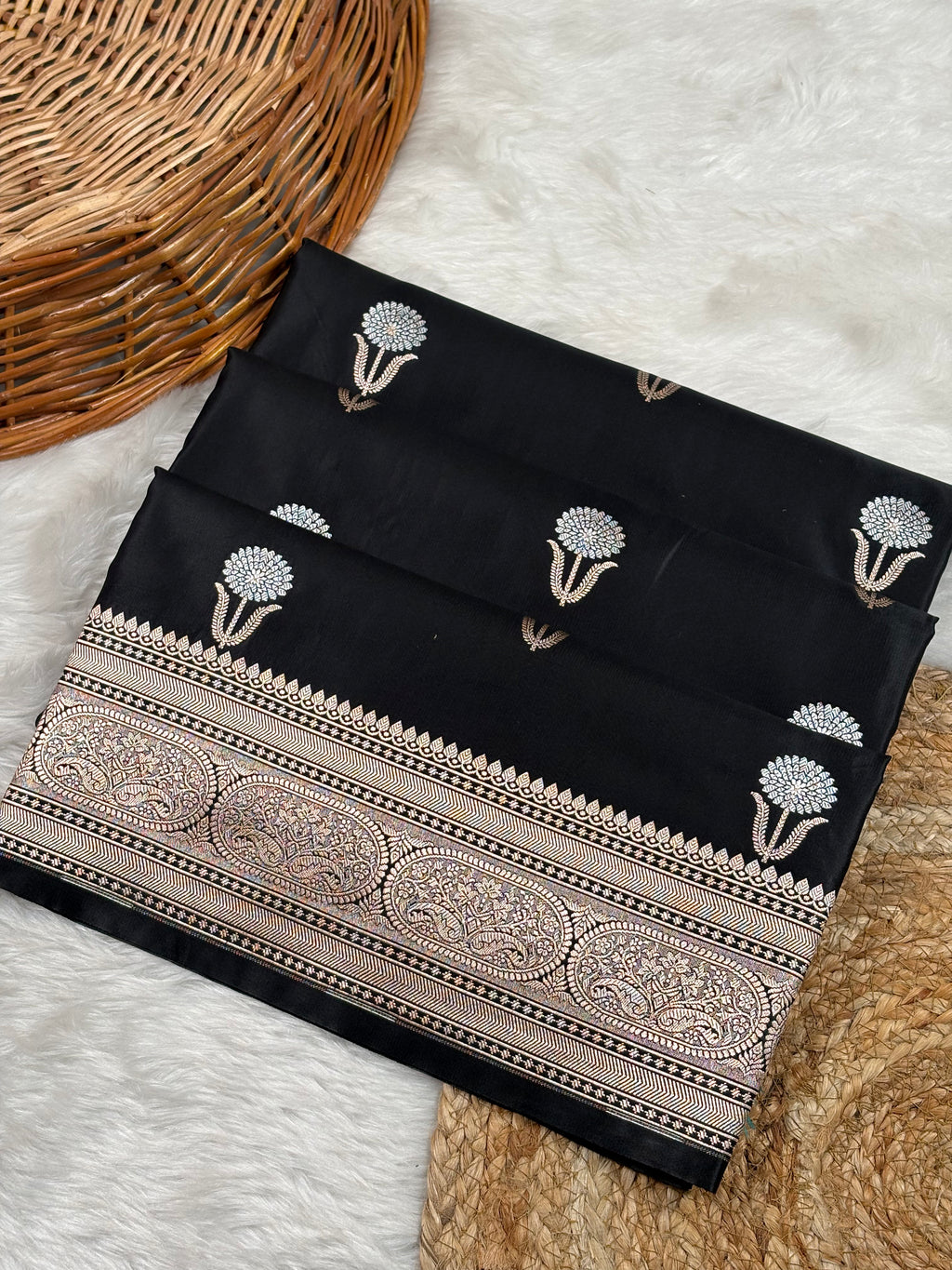 Black mashru katan banarasi saree with sona rupa floral motifs and a wide ornate paisley border, laid flat on a white faux-fur surface with a wicker basket and woven mat as props.