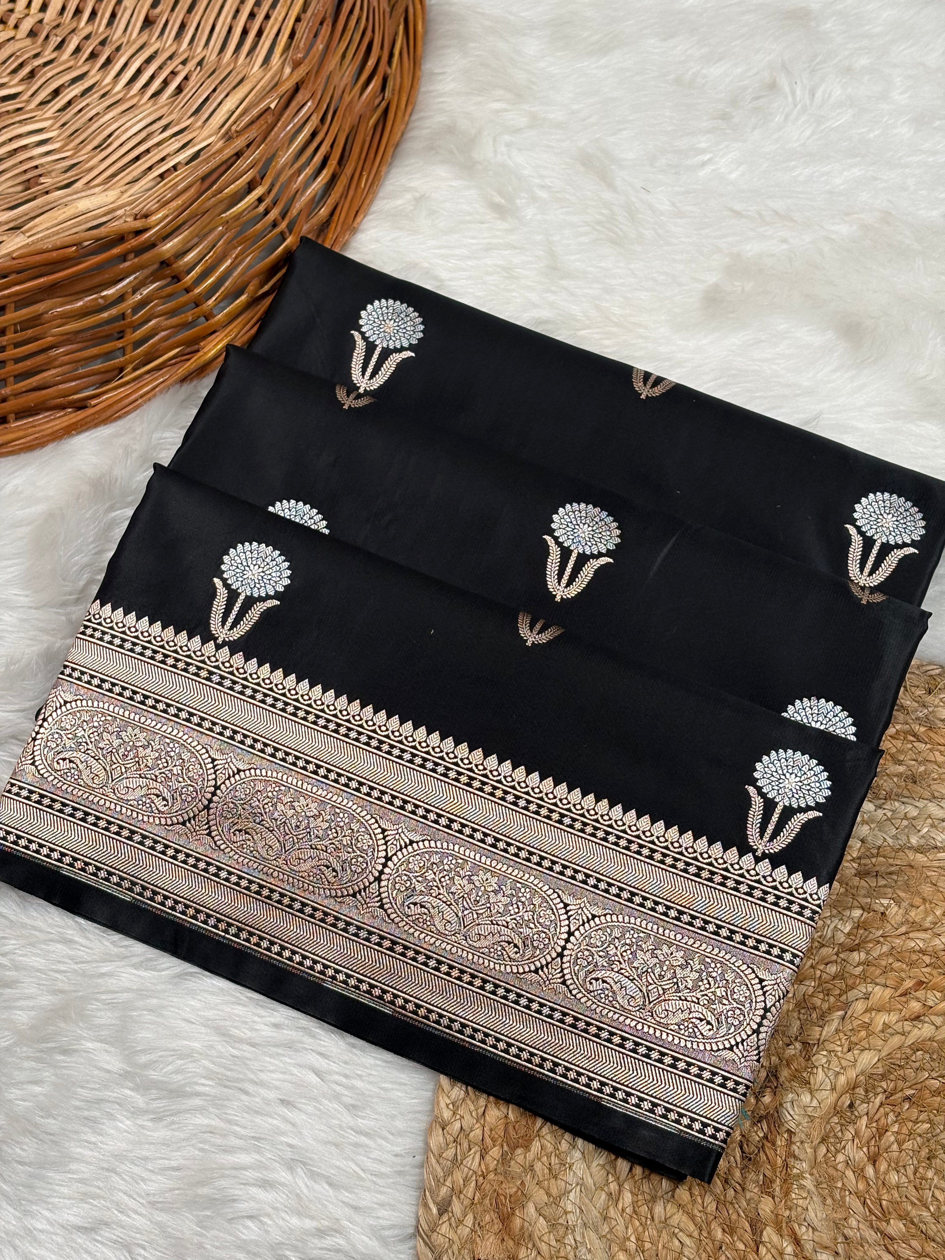 Black mashru katan banarasi saree with sona rupa floral motifs and a wide ornate paisley border, laid flat on a white faux-fur surface with a wicker basket and woven mat as props.