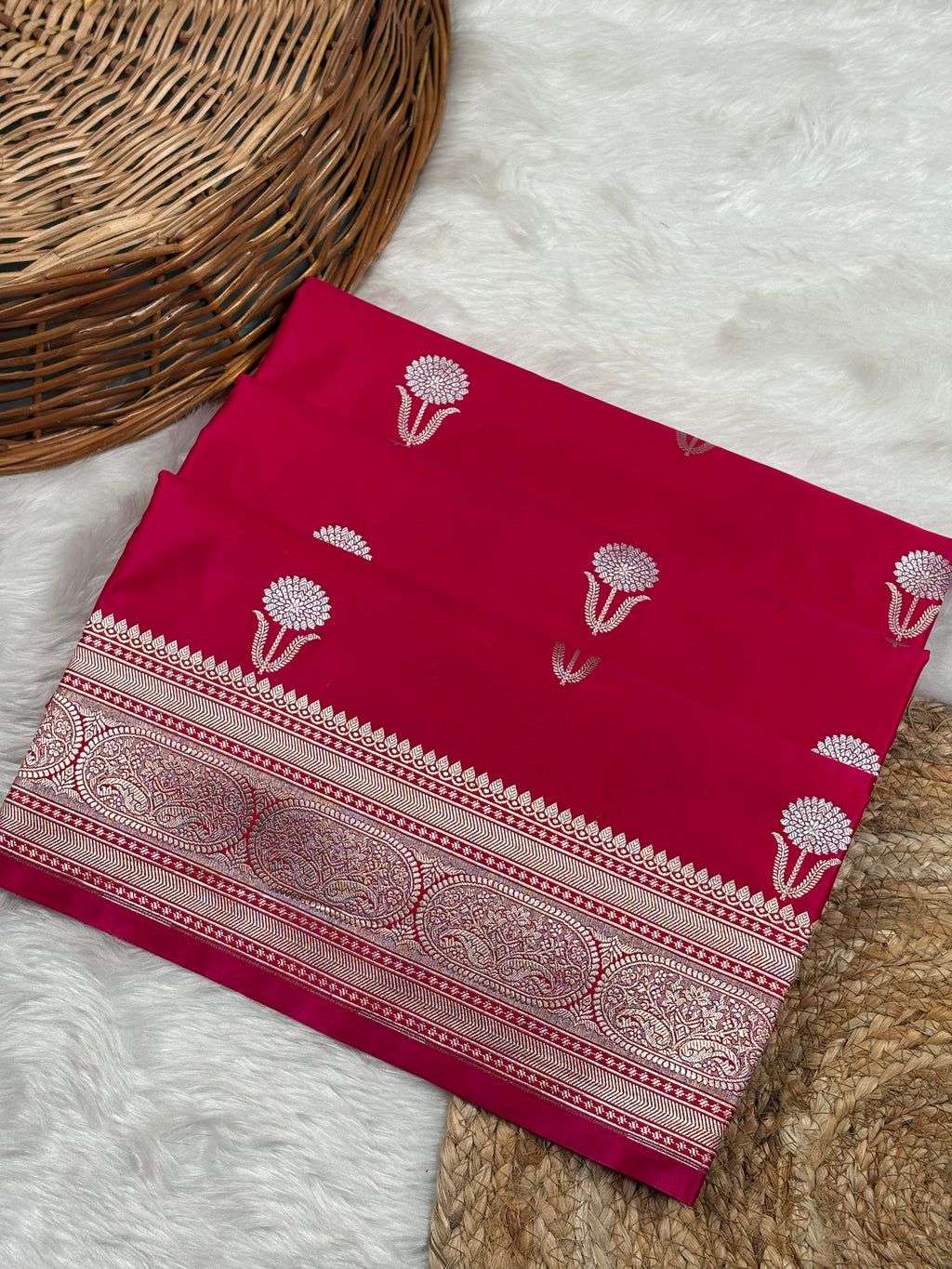 A folded deep magenta pink saree with sona rupa floral weaving and border motifs, displayed on a white faux fur surface next to a woven wicker basket.