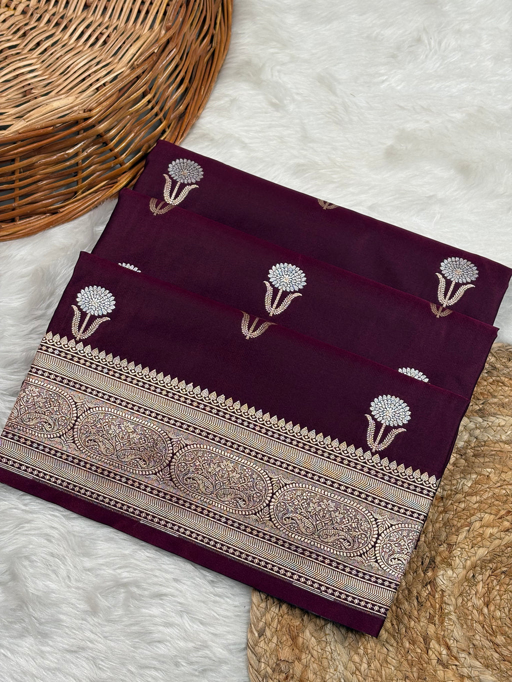 A folded deep maroon silk saree with sona rupa floral weaving, displayed on a white faux fur rug next to a wicker basket.