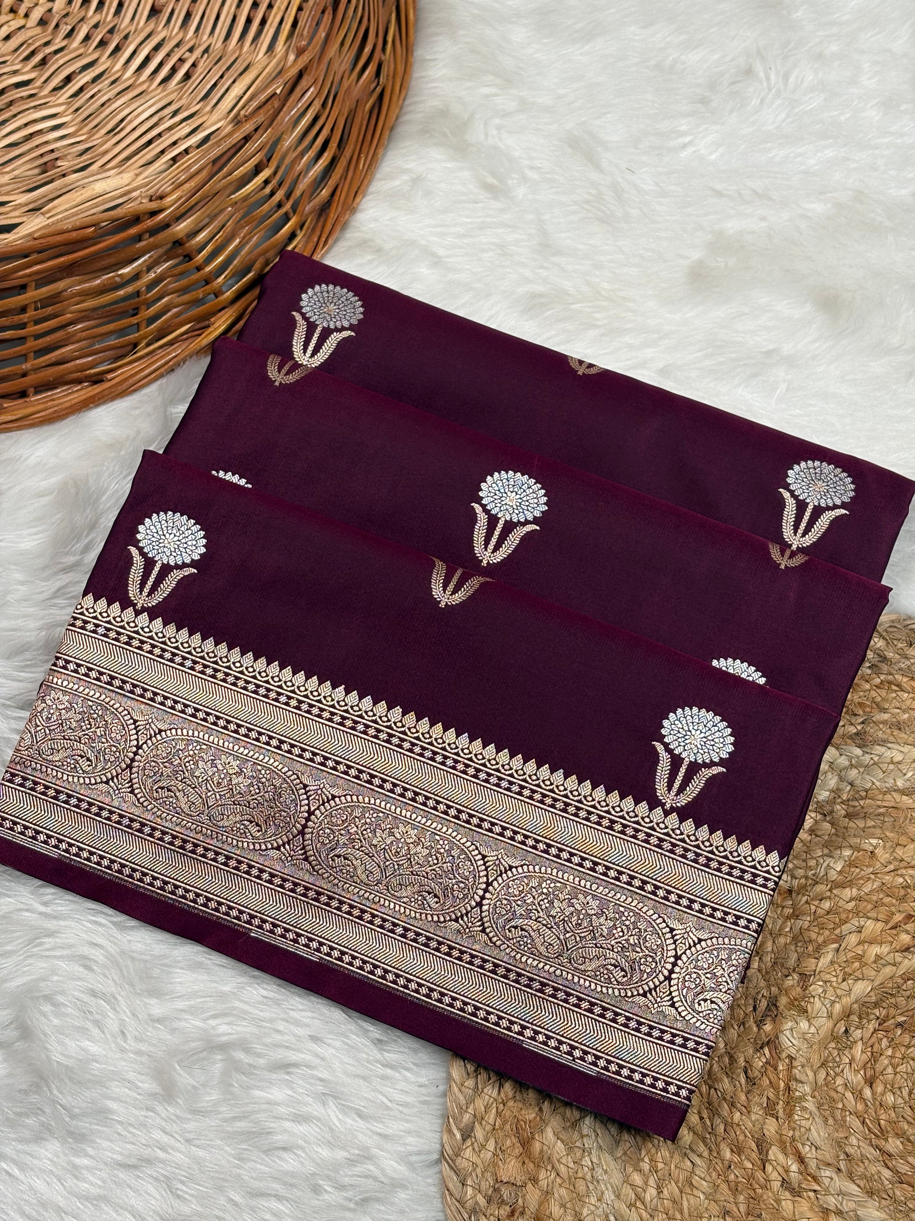 A folded deep maroon silk saree with sona rupa floral weaving, displayed on a white faux fur rug next to a wicker basket.