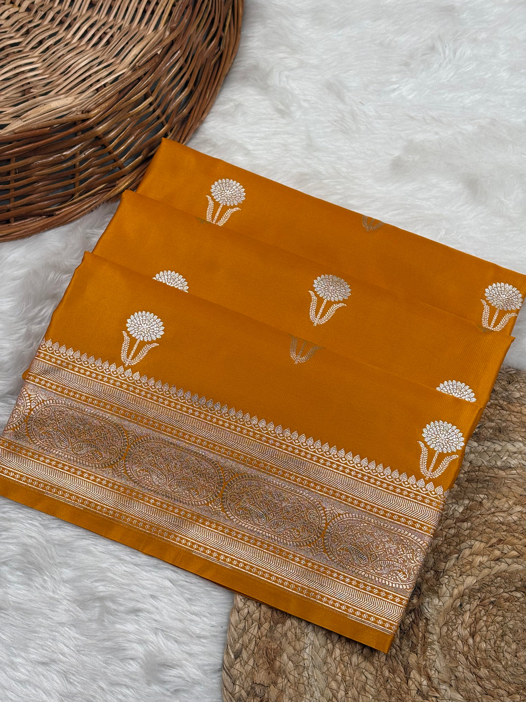 A folded mustard yellow Banarasi silk saree with sona rupa motifs and a gold border, displayed on a white faux fur rug next to a woven wicker basket.
