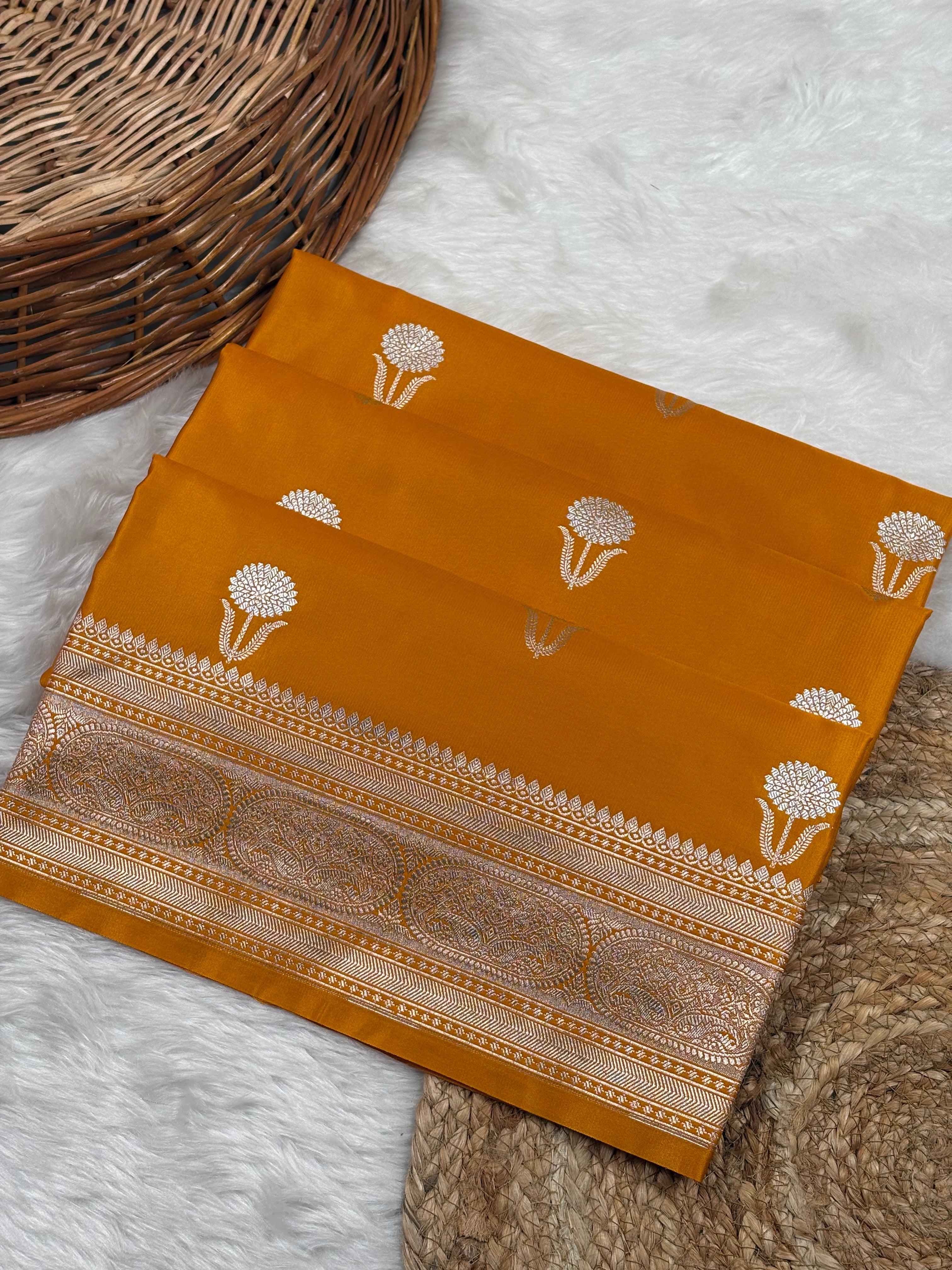 A folded mustard yellow Banarasi silk saree with sona rupa motifs and a gold border, displayed on a white faux fur rug next to a woven wicker basket.