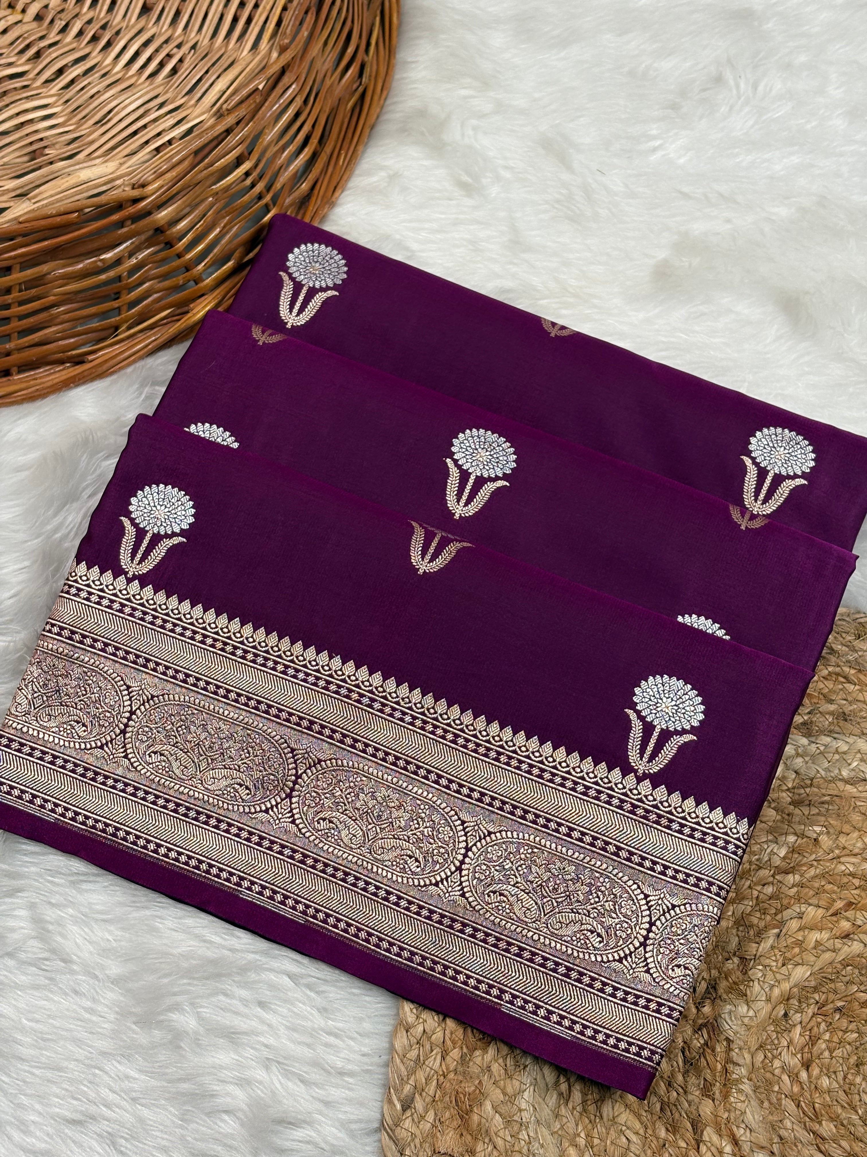 A folded deep purple saree, featuring sona rupa floral motifs and a detailed gold border, styled on a white faux fur surface next to a wicker basket.