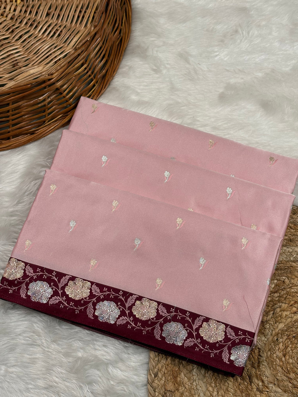 A folded dusty pink saree, featuring small zari weaving motifs and a contrasting deep maroon border with floral sona rupa zari work, displayed on a white faux fur rug next to a wicker basket.