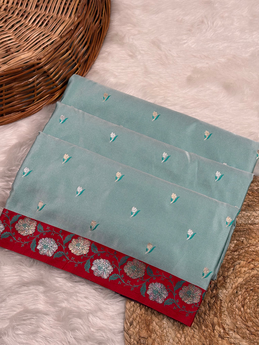 A folded sage green mashru katan saree, featuring small floral motifs and a contrasting red border with a floral weaving, styled on a white faux fur rug next to a wicker basket.