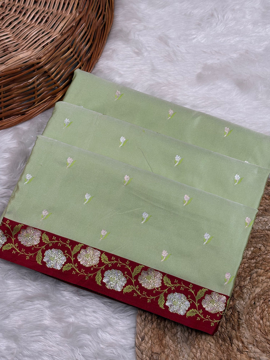 A folded light green saree with small floral motifs and a rich contrast red border featuring white and green floral weaving, displayed on a white faux fur surface next to a wicker basket.