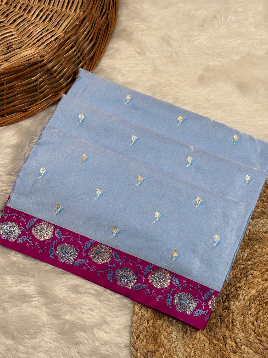 A folded light blue saree with small motifs and a rich magenta border featuring zari floral patterns, displayed on a white faux fur surface next to a wicker basket.