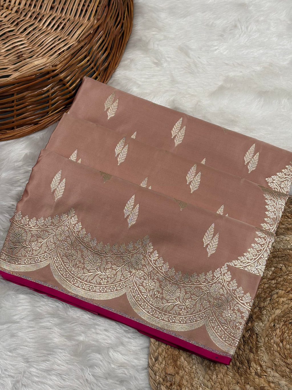 A folded dusty rose pink saree with zari leaf and border patterns, displayed on a white faux fur rug next to a wicker basket.