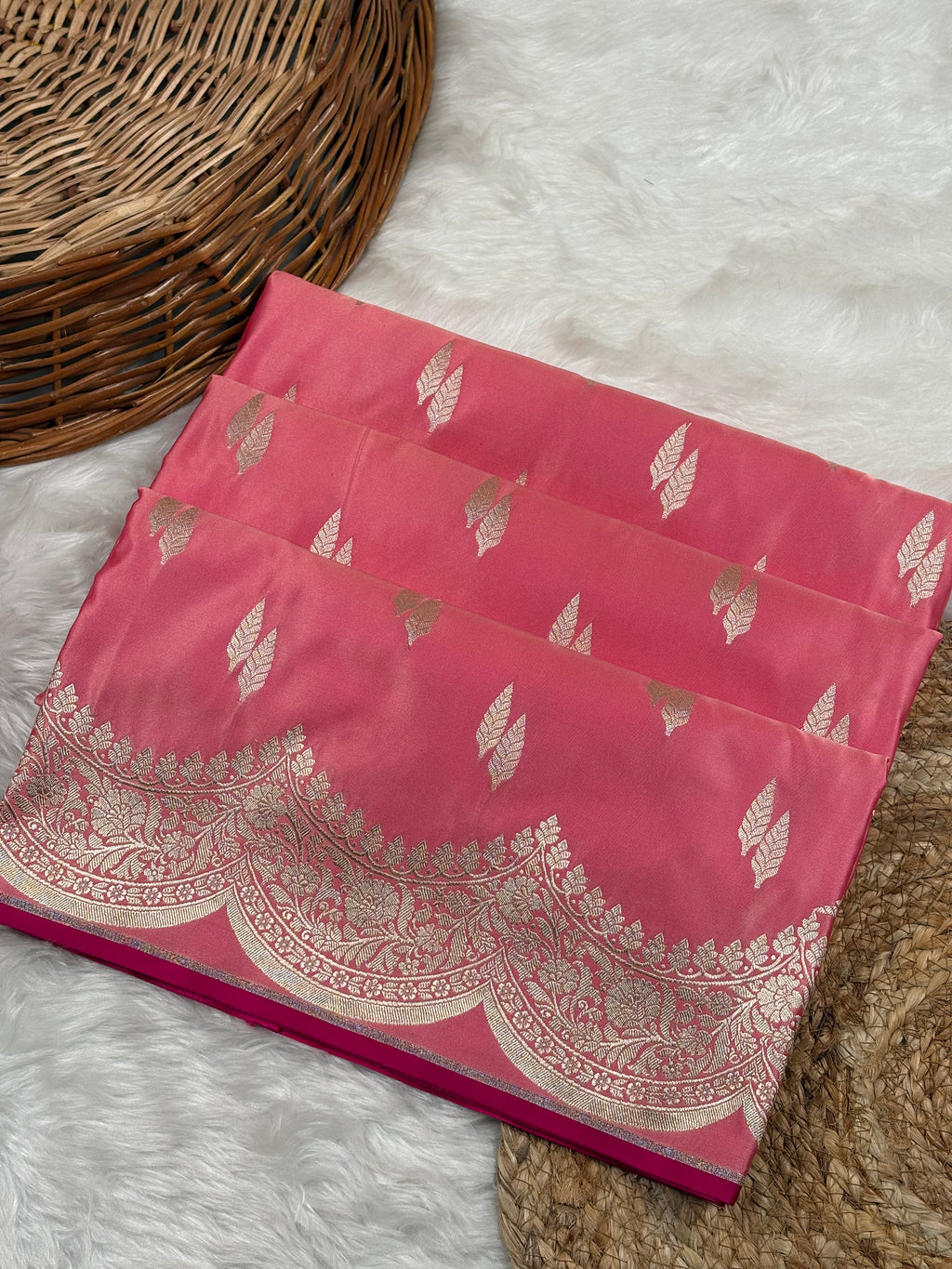 A folded pink silk saree with gold zari leaf and border motifs, displayed on a white faux fur rug next to a woven wicker basket.