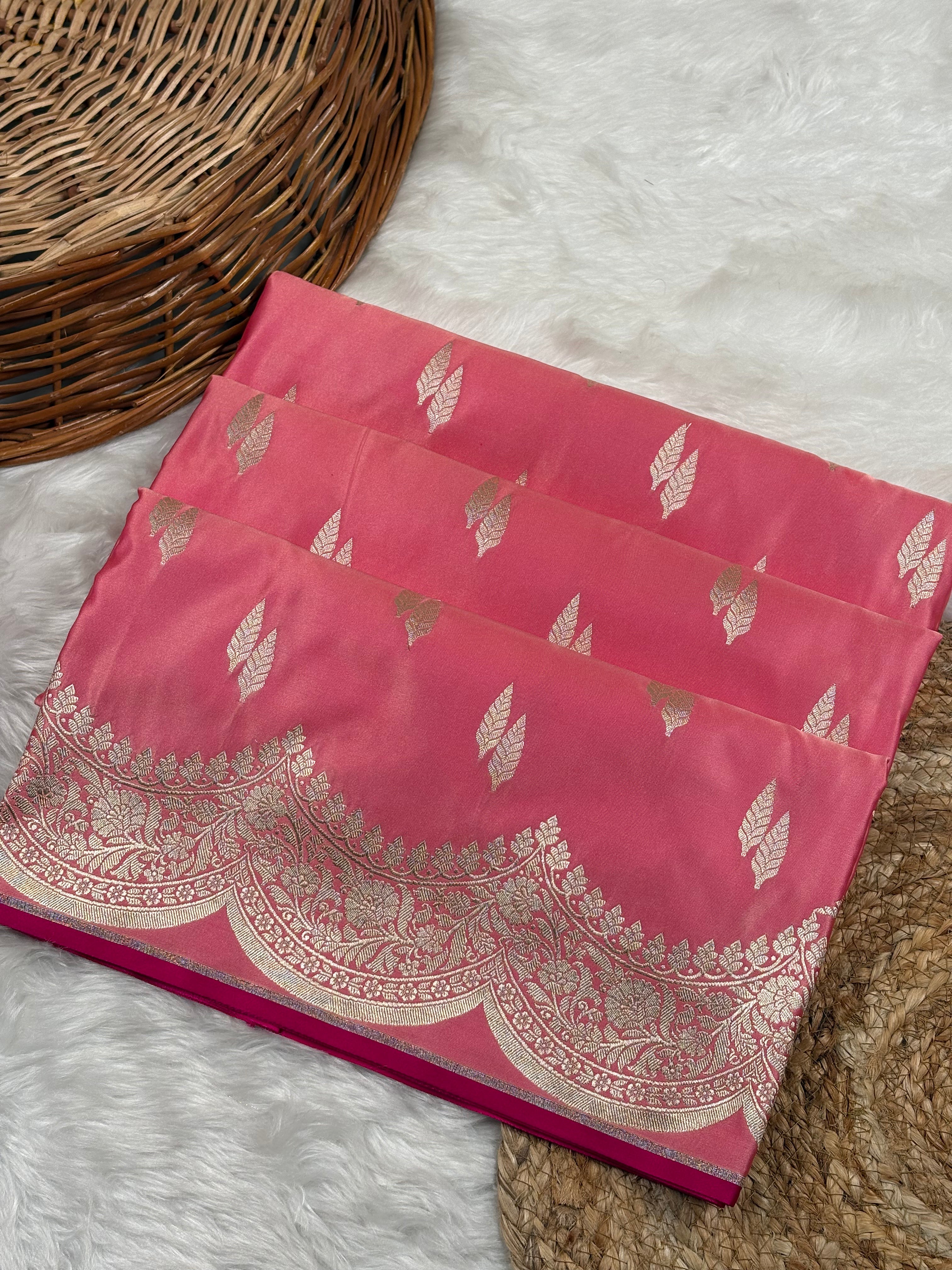 A folded pink silk saree with gold zari leaf and border motifs, displayed on a white faux fur rug next to a woven wicker basket.