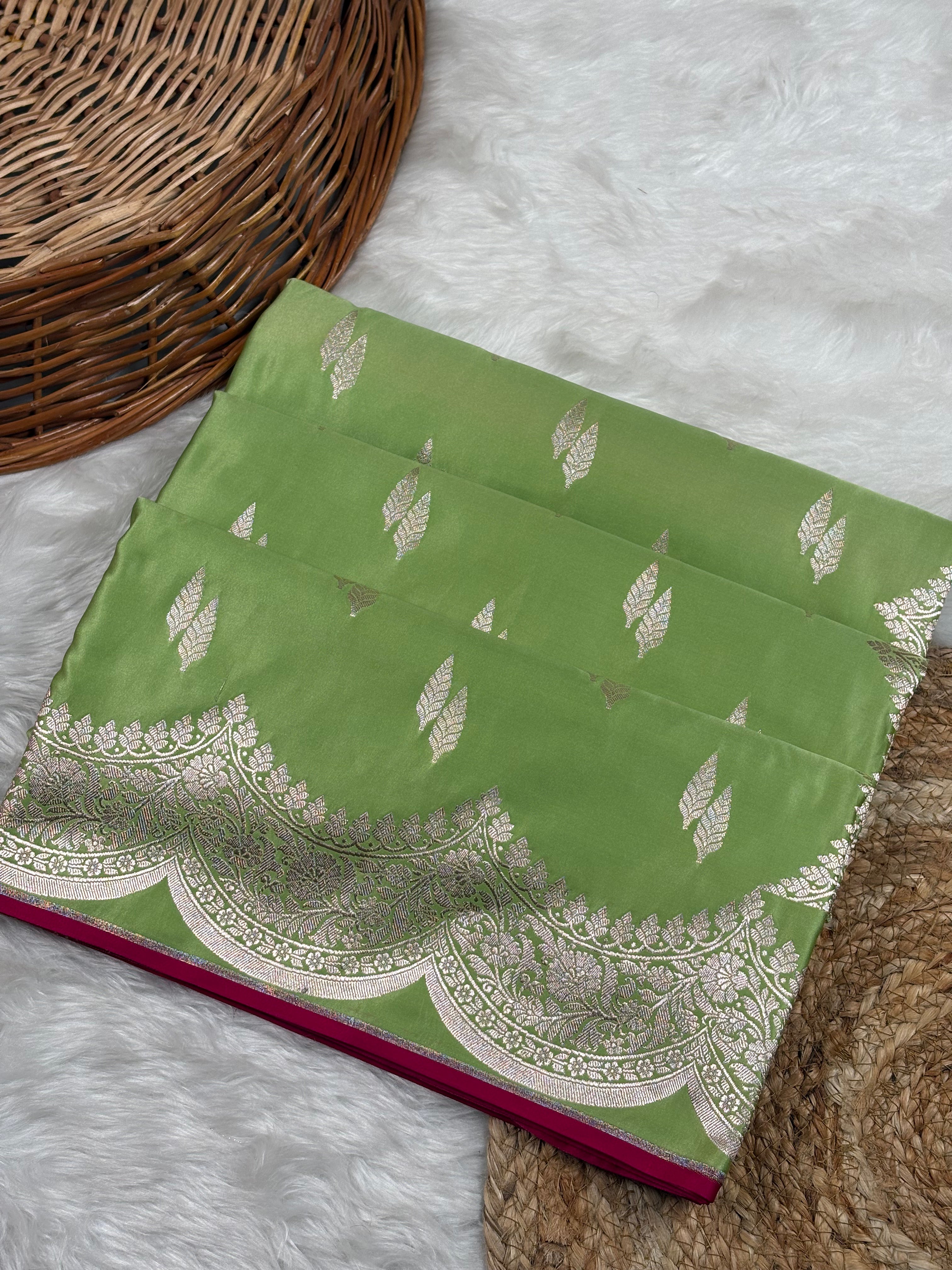 A folded sage green saree with zari leaf motifs and a scalloped border, displayed on a white faux fur surface next to a wicker basket.