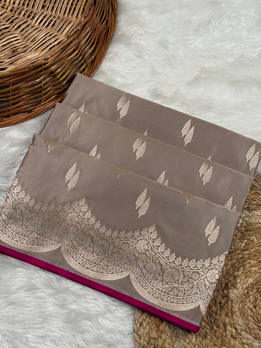 A folded taupe-brown saree with gold zari leaf motifs and a scalloped gold border, displayed on a white faux fur rug next to a wicker basket.