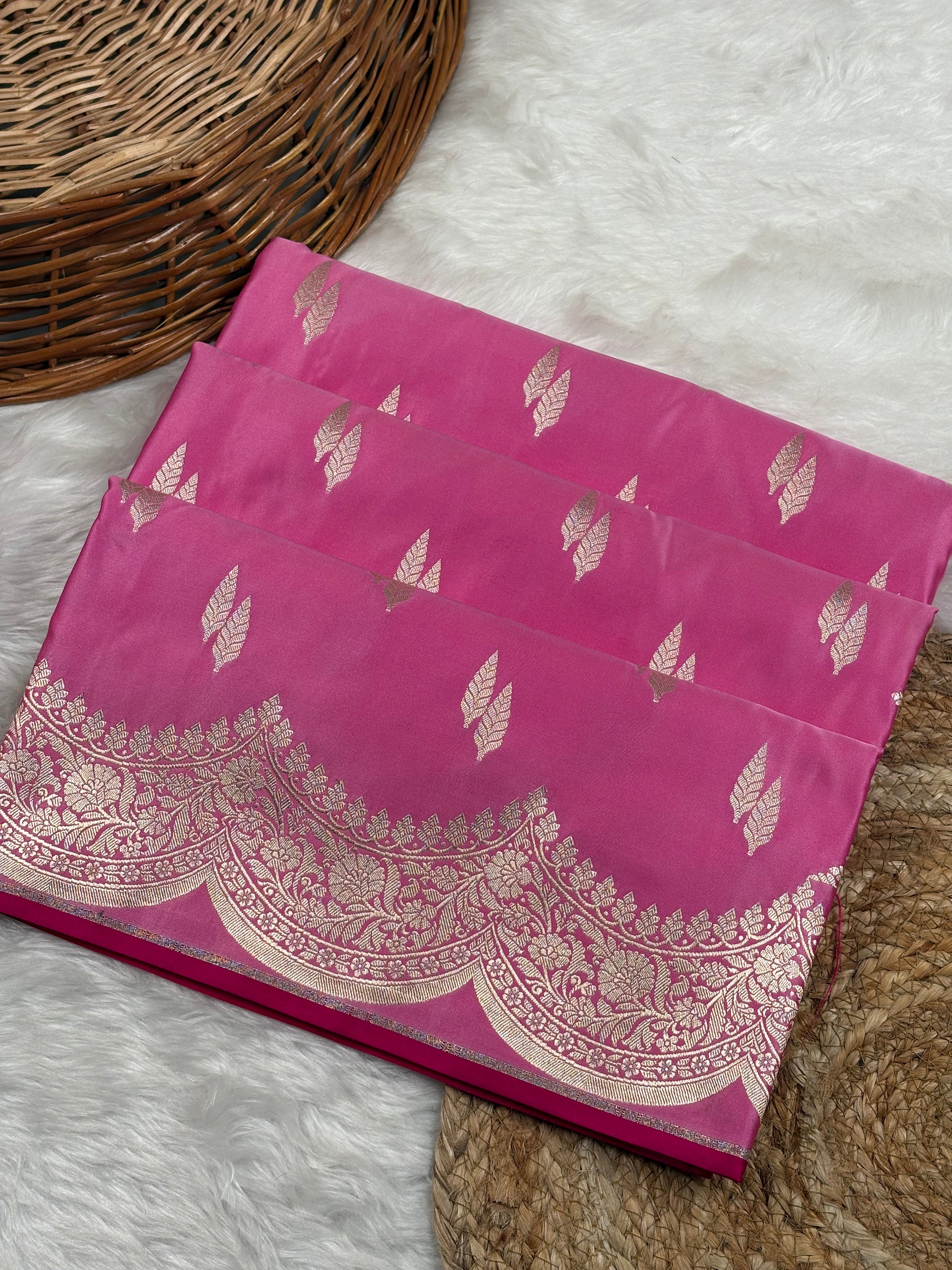 A folded pink silk saree with zari motifs and a decorative scalloped border, displayed on a white faux fur surface next to a wicker basket.