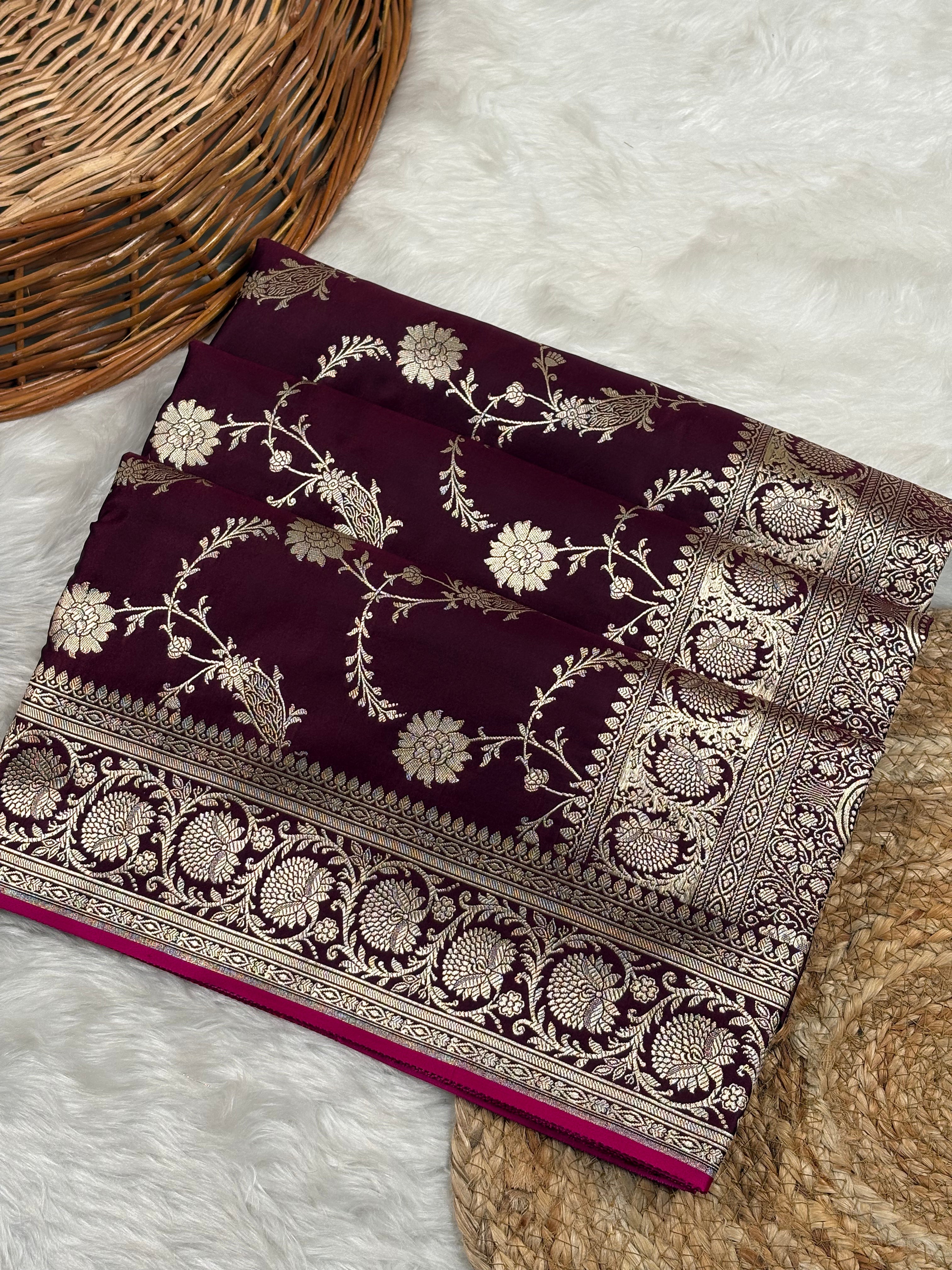 A folded deep maroon silk saree with intricate gold zari floral motifs and borders weaving, displayed on a white faux fur surface next to a woven wicker basket.