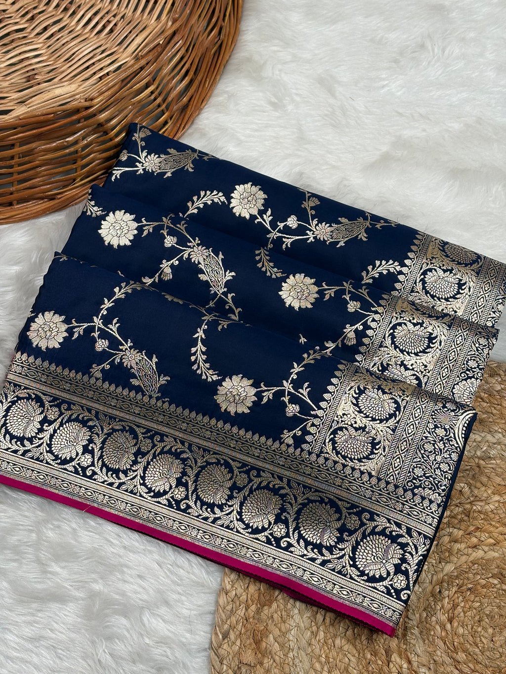 A folded navy blue Banarasi silk saree with intricate zari floral weaving motifs, displayed on a white faux fur rug next to a wicker basket.