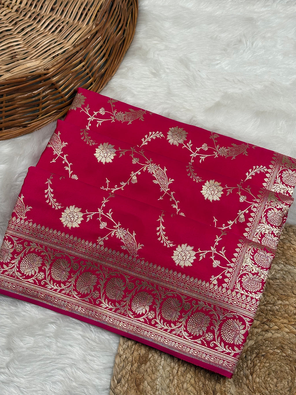 A folded raani pink saree with intricate gold floral motifs weaving, displayed on a white faux fur surface next to a woven wicker basket.