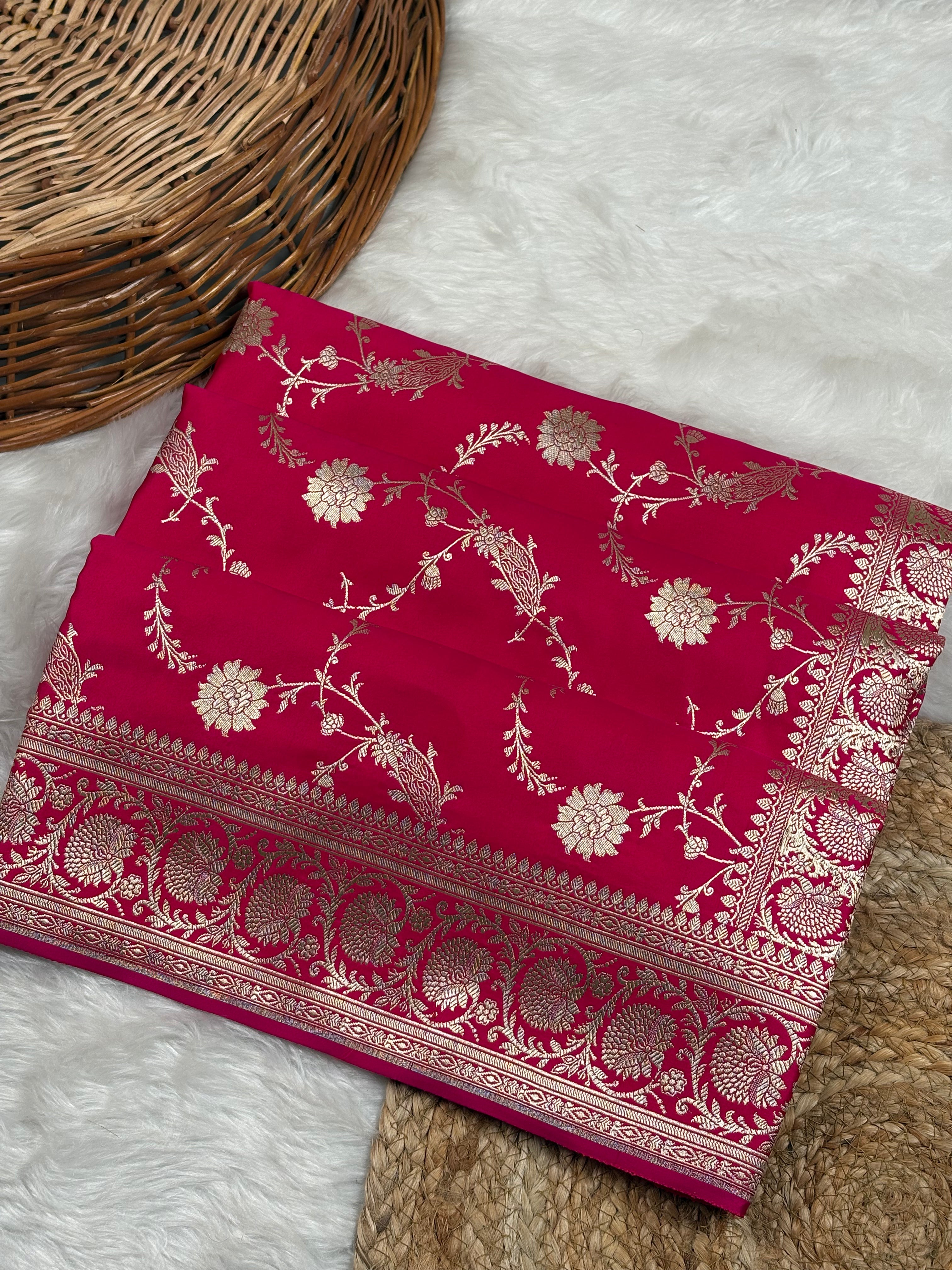 A folded raani pink saree with intricate gold floral motifs weaving, displayed on a white faux fur surface next to a woven wicker basket.