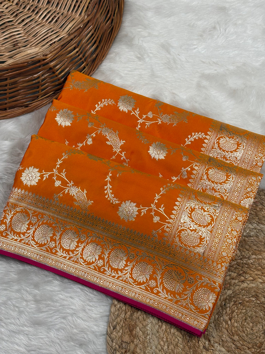 An orange Banarasi silk saree with intricate gold zari floral weaving, folded neatly on a white faux fur surface next to a woven wicker basket.