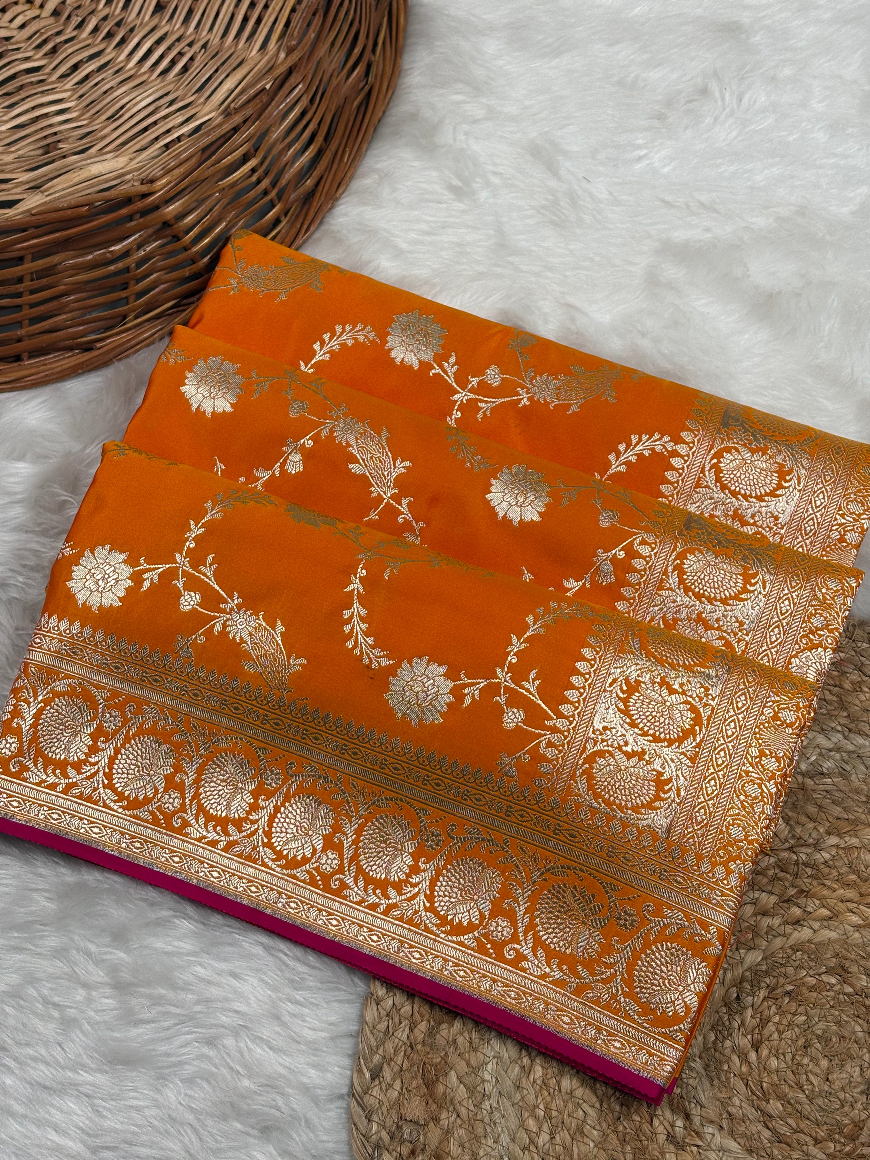 An orange Banarasi silk saree with intricate gold zari floral weaving, folded neatly on a white faux fur surface next to a woven wicker basket.