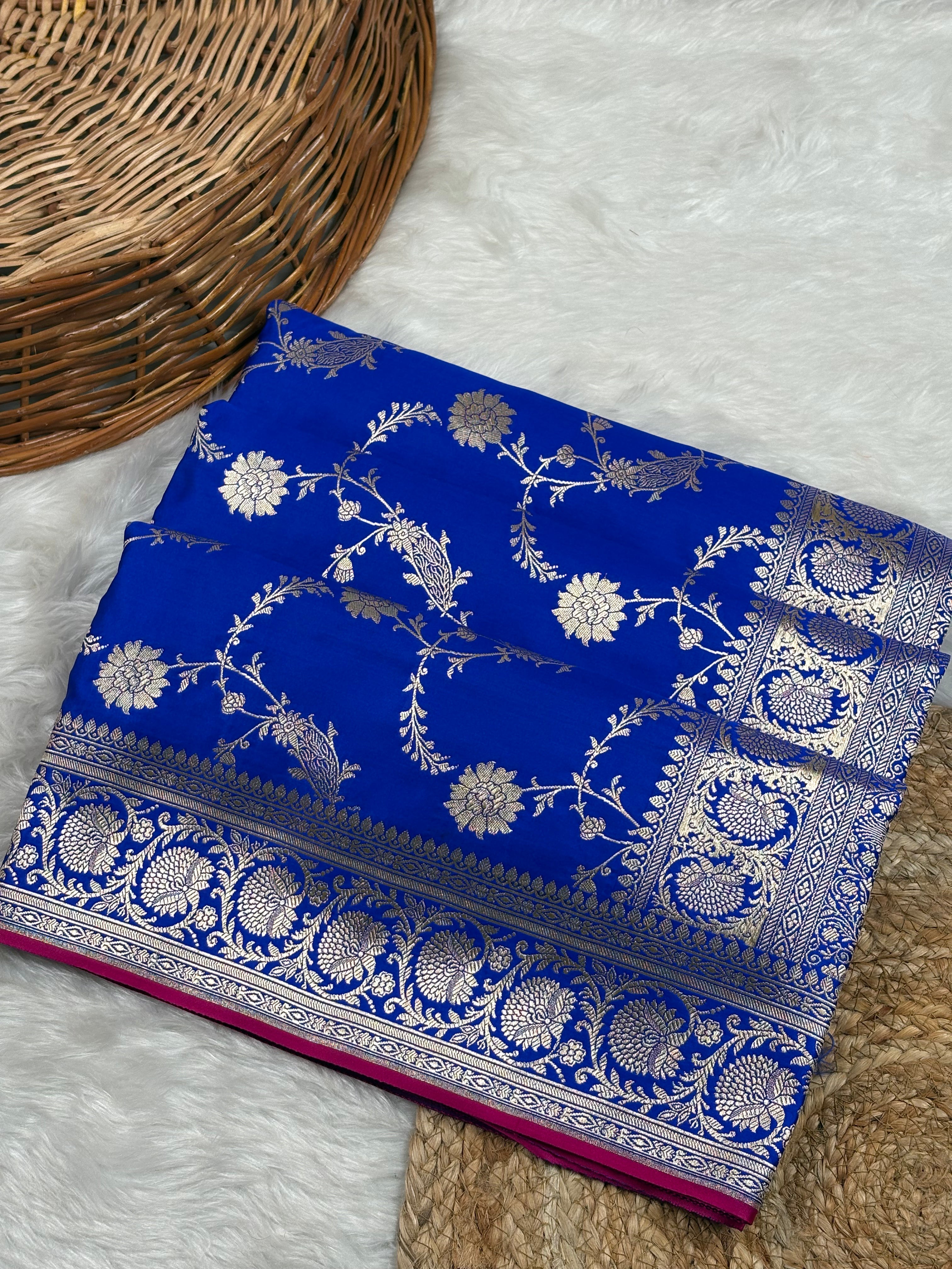 A folded royal blue silk saree with intricate gold zari floral motifs, displayed on a white faux fur surface next to a woven wicker basket.