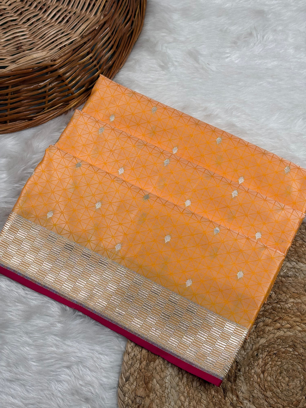 An orange Banarasi silk saree with a gold geometric pattern and a gold zari border, displayed on a white faux fur rug next to a wicker basket.