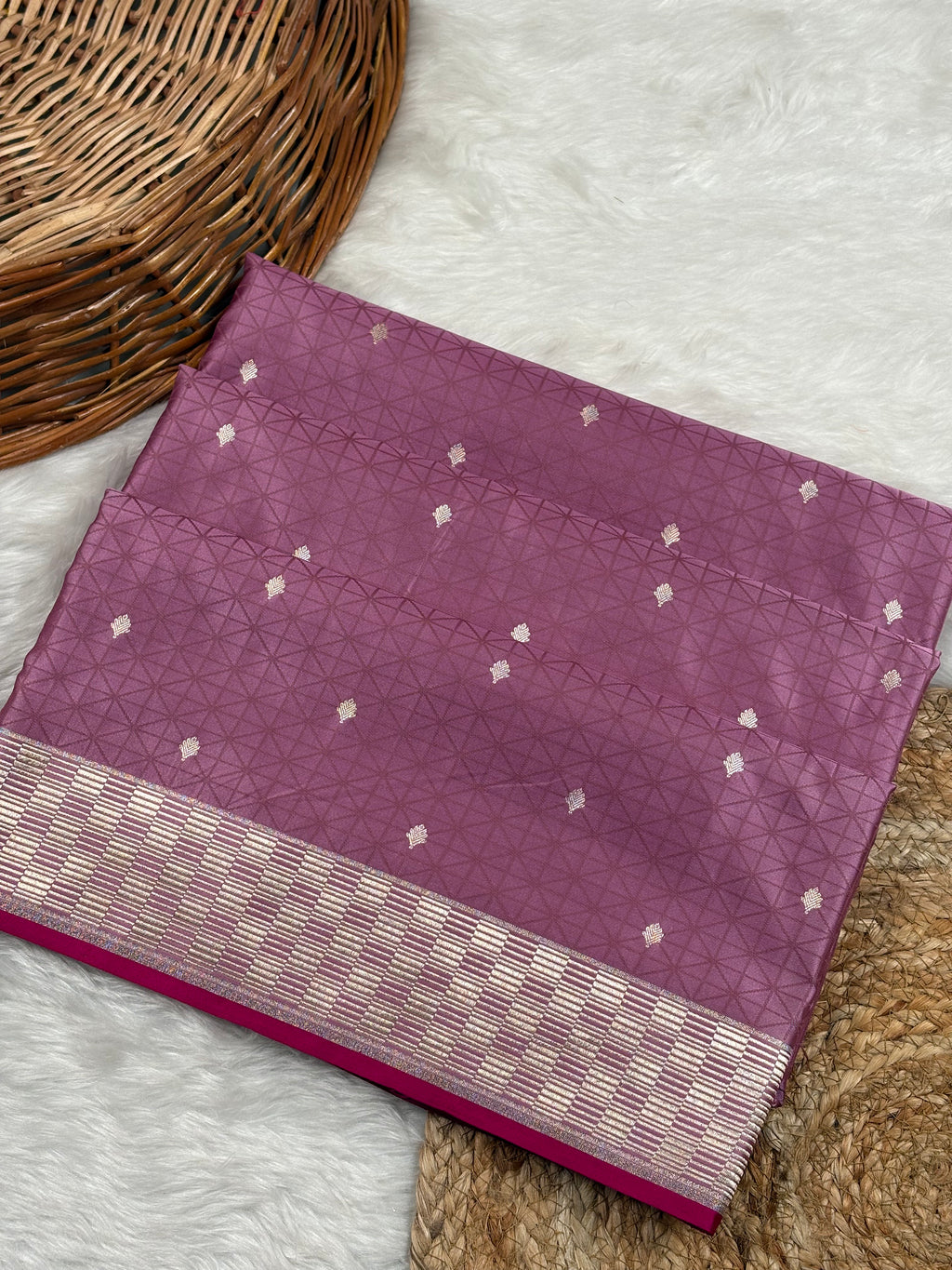 A folded purple silk saree with a tanchui geometric pattern and a gold zari weaving border, displayed on a white faux fur rug next to a woven wicker basket.