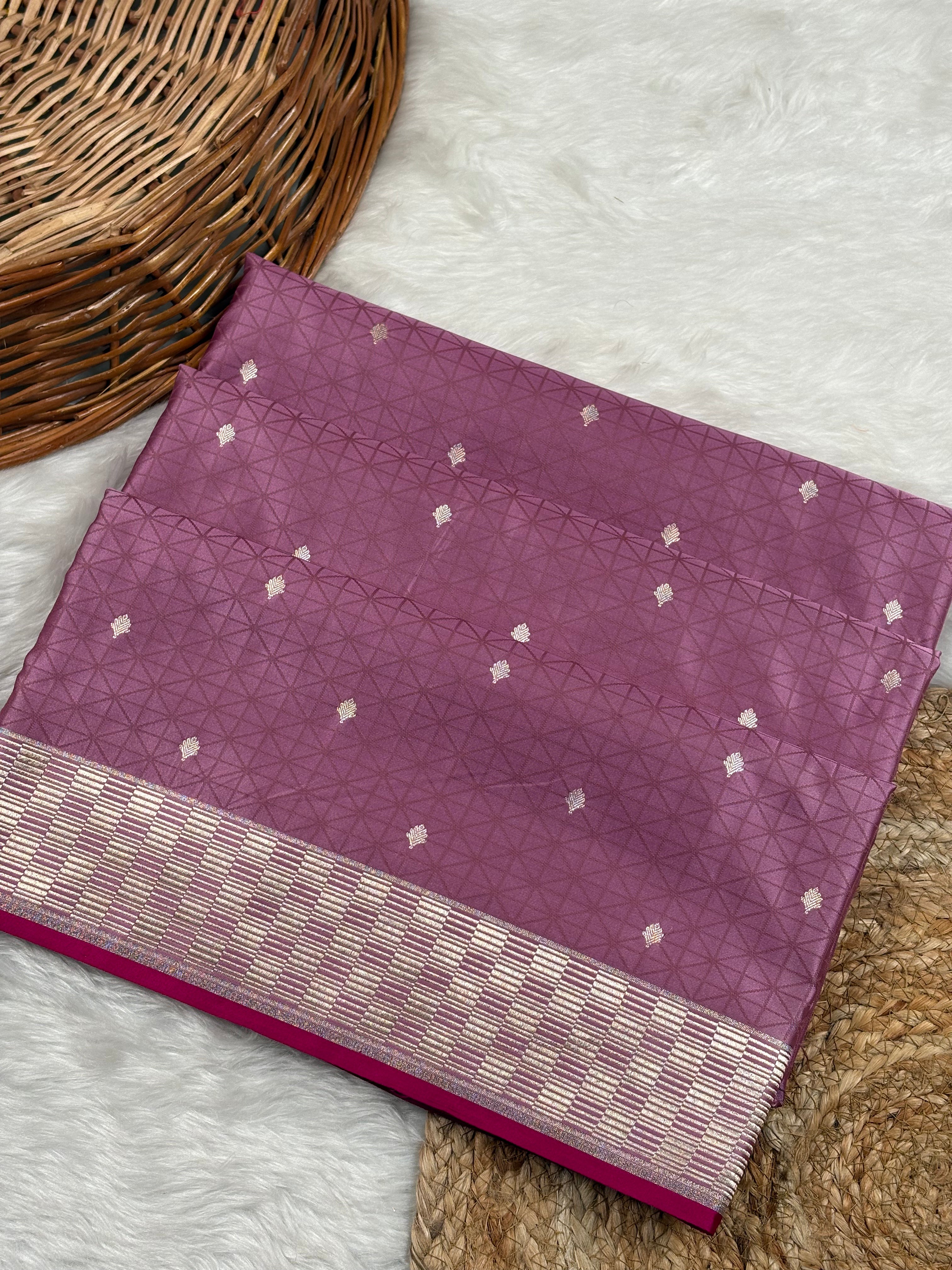 A folded purple silk saree with a tanchui geometric pattern and a gold zari weaving border, displayed on a white faux fur rug next to a woven wicker basket.