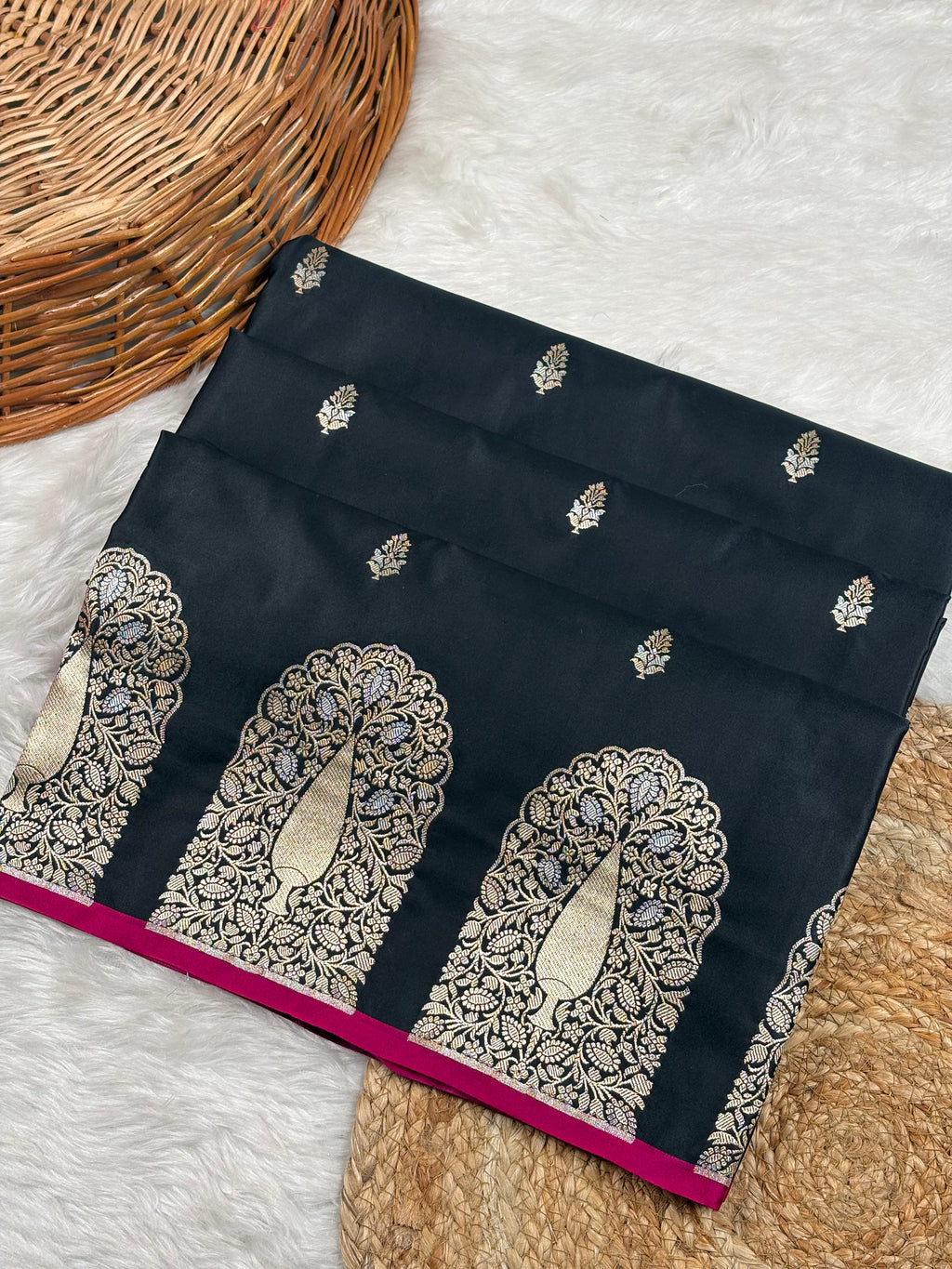 A folded black mashru katan saree with ornate alfy mina and floral border weaving, displayed on a white faux fur surface with a wicker basket and woven mat as props.